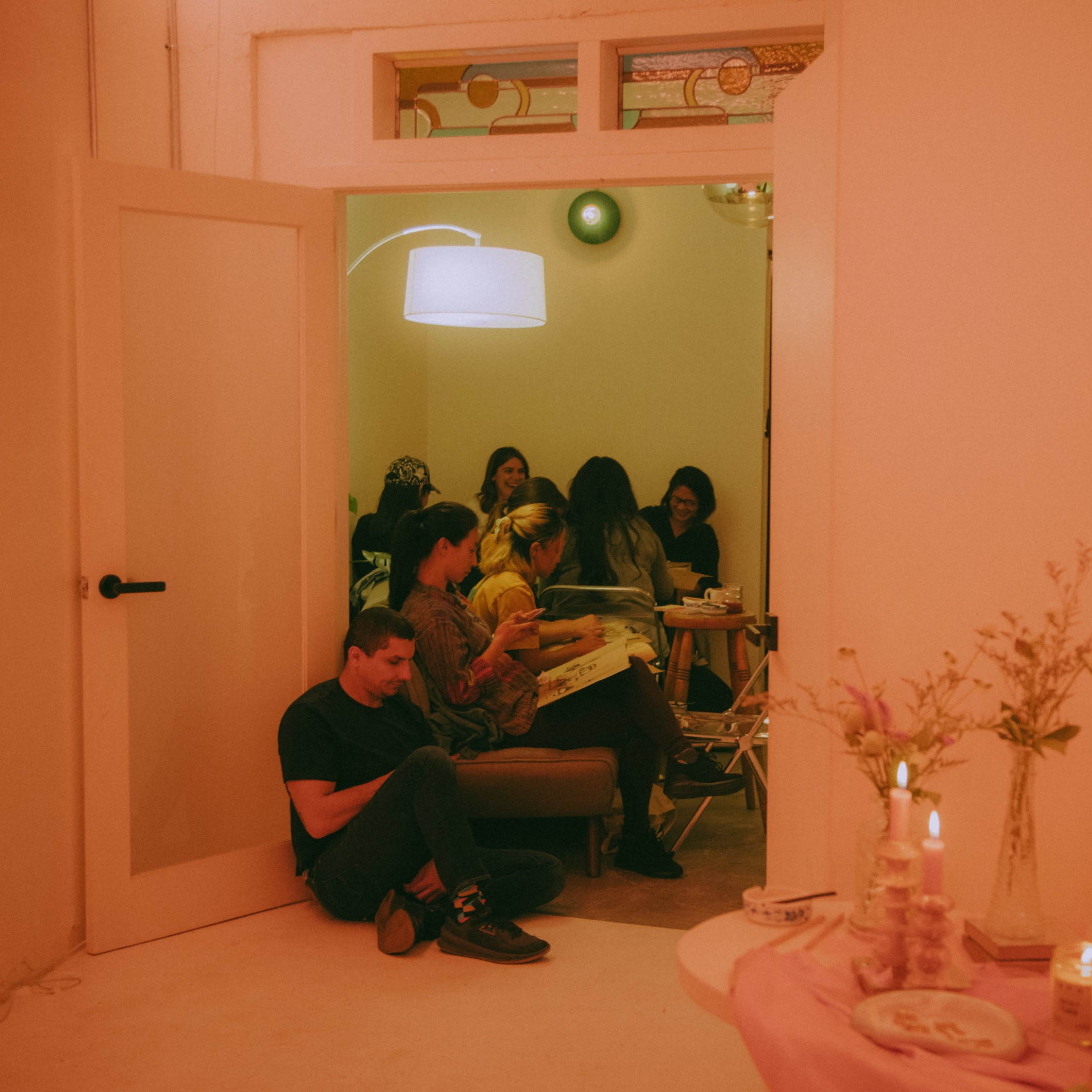 A group of people gathers around a table in a brightly lit room while another person sits on the floor near the doorway.