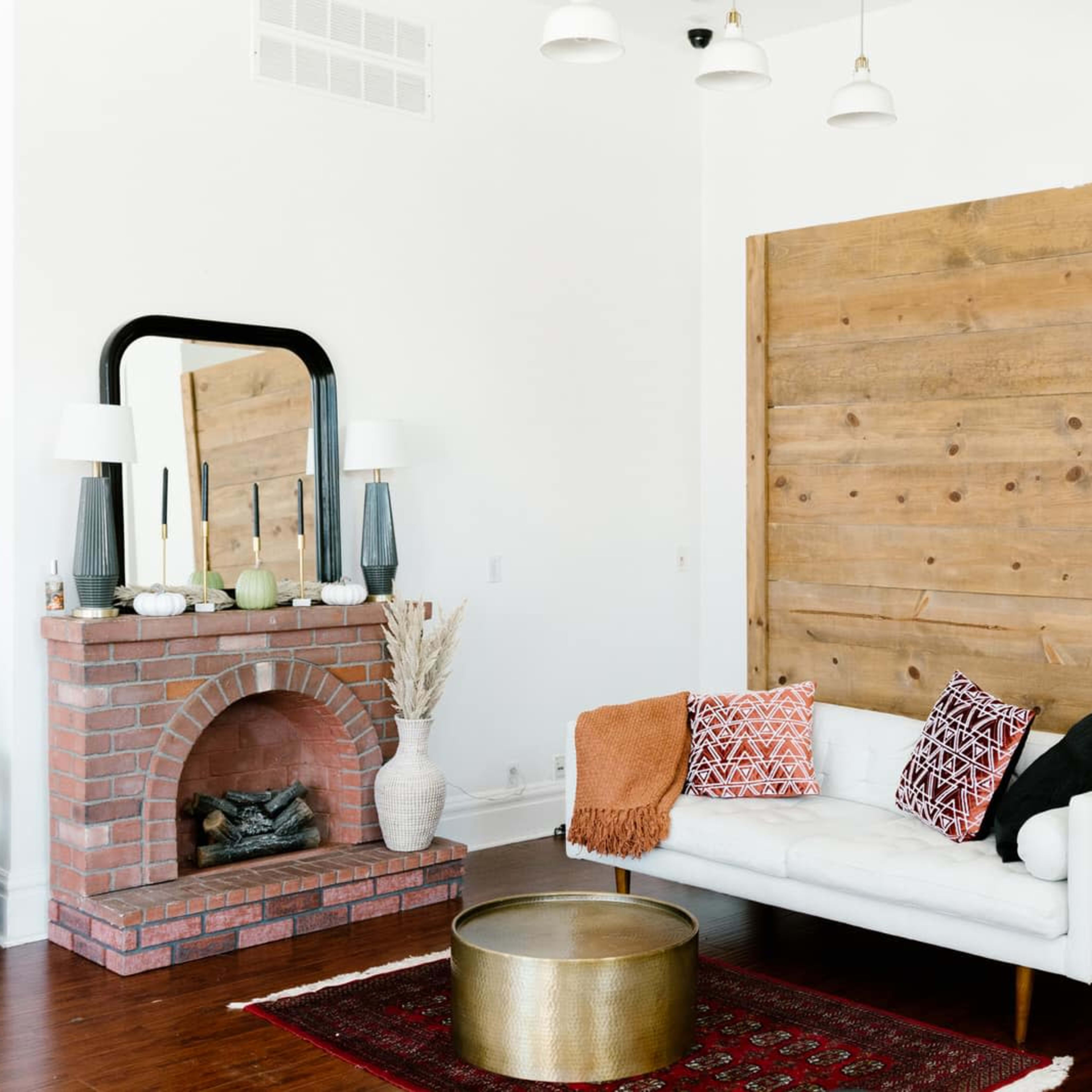 The living room features a white sofa with patterned pillows, a brick fireplace beneath a mirror, a wooden accent wall, and a potted plant beside a round coffee table.