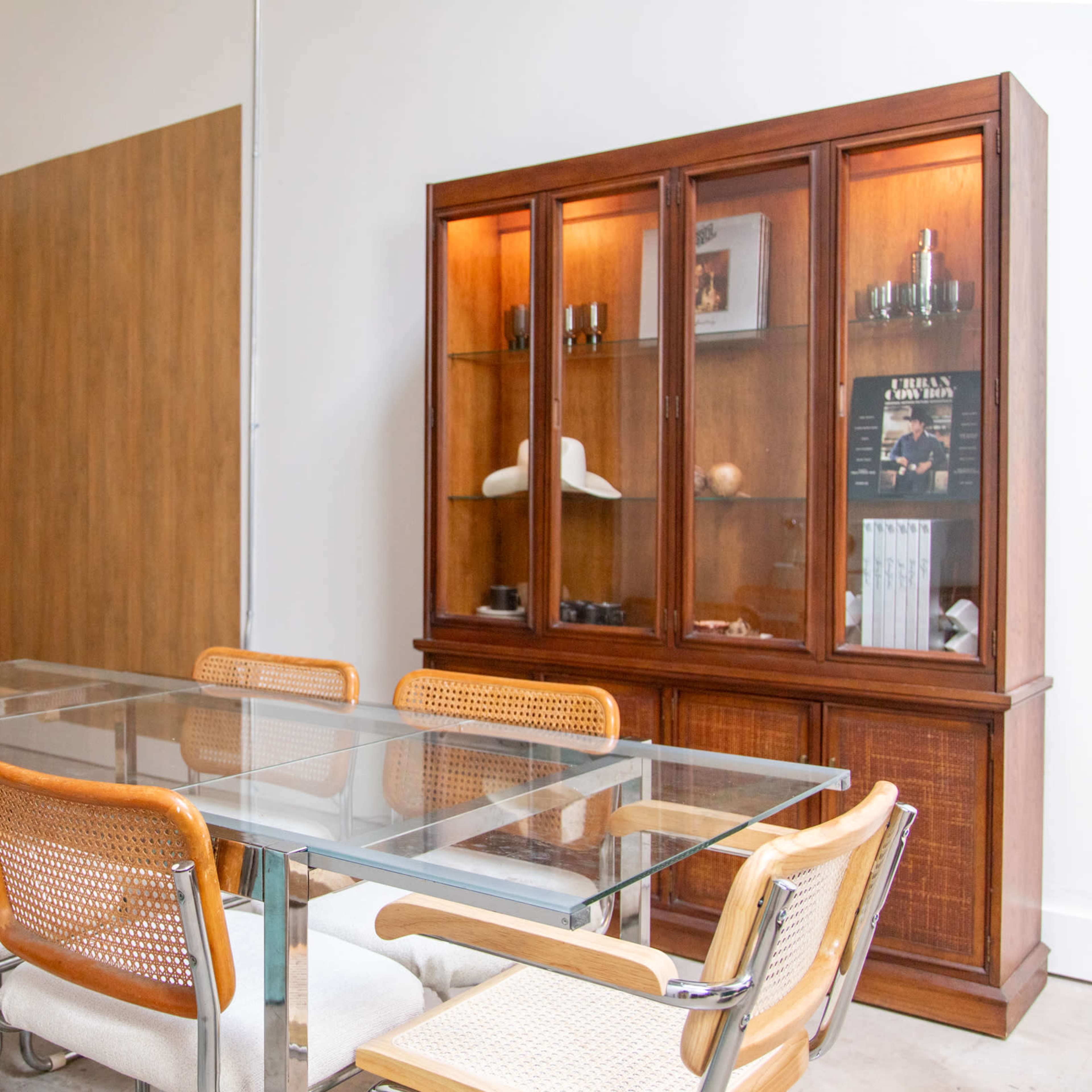 A modern dining area features a glass table surrounded by rattan chairs, with a wooden display cabinet and a wall panel in the background.