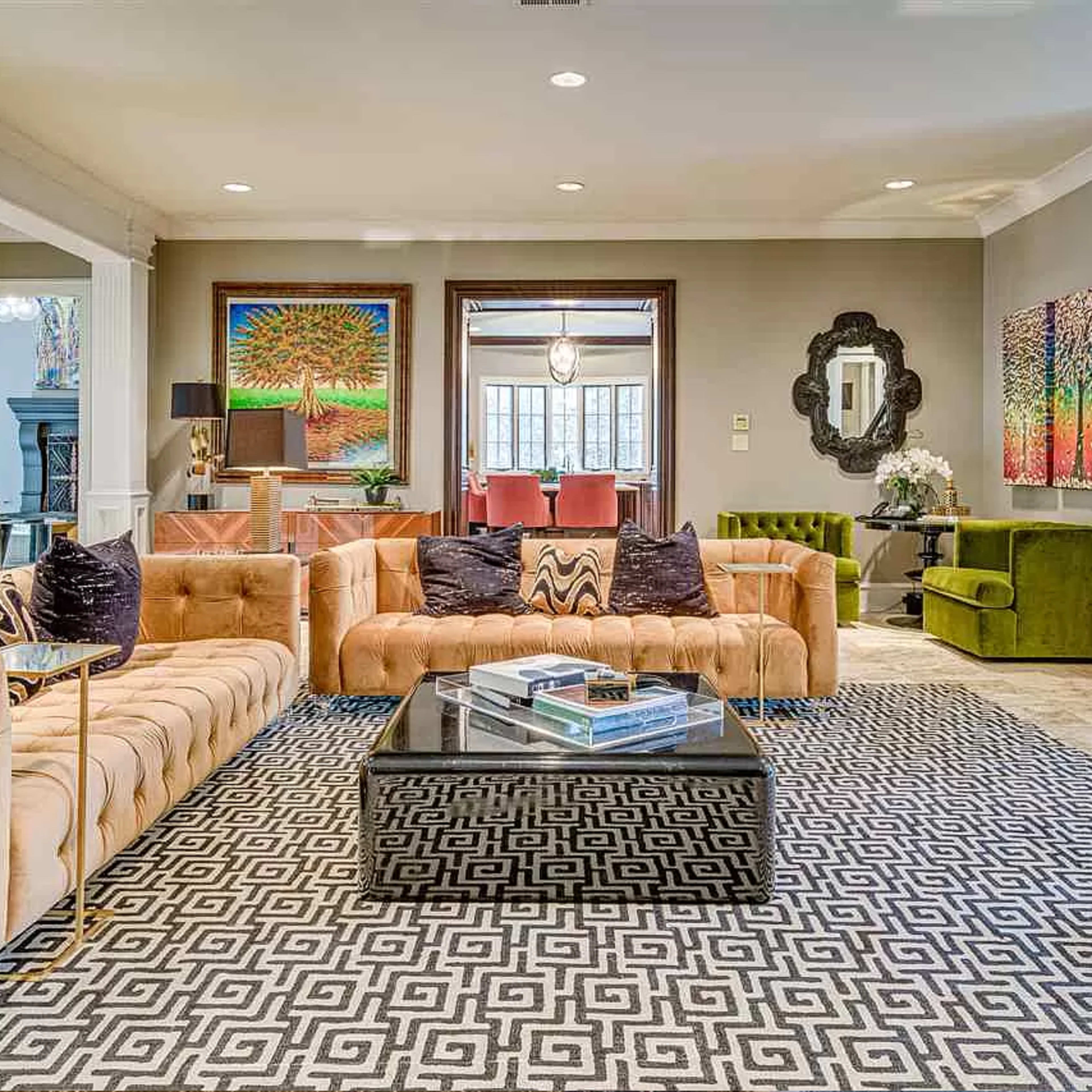 The living room features a large, beige tufted sofa, colorful wall art, a dark coffee table, and a patterned area rug, with a staircase visible in the background.