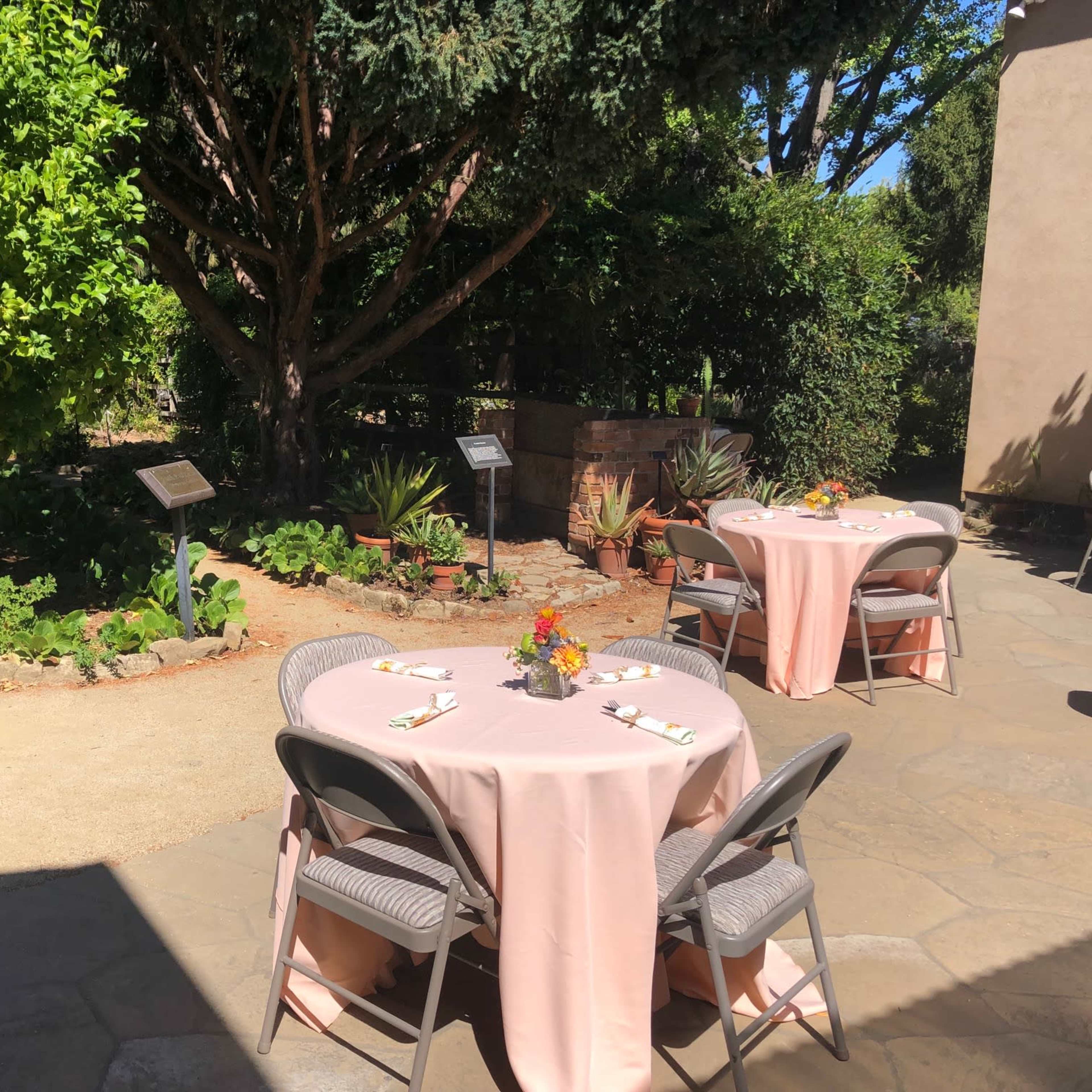 The scene features two round tables set with pink tablecloths and floral centerpieces in a garden area surrounded by greenery.