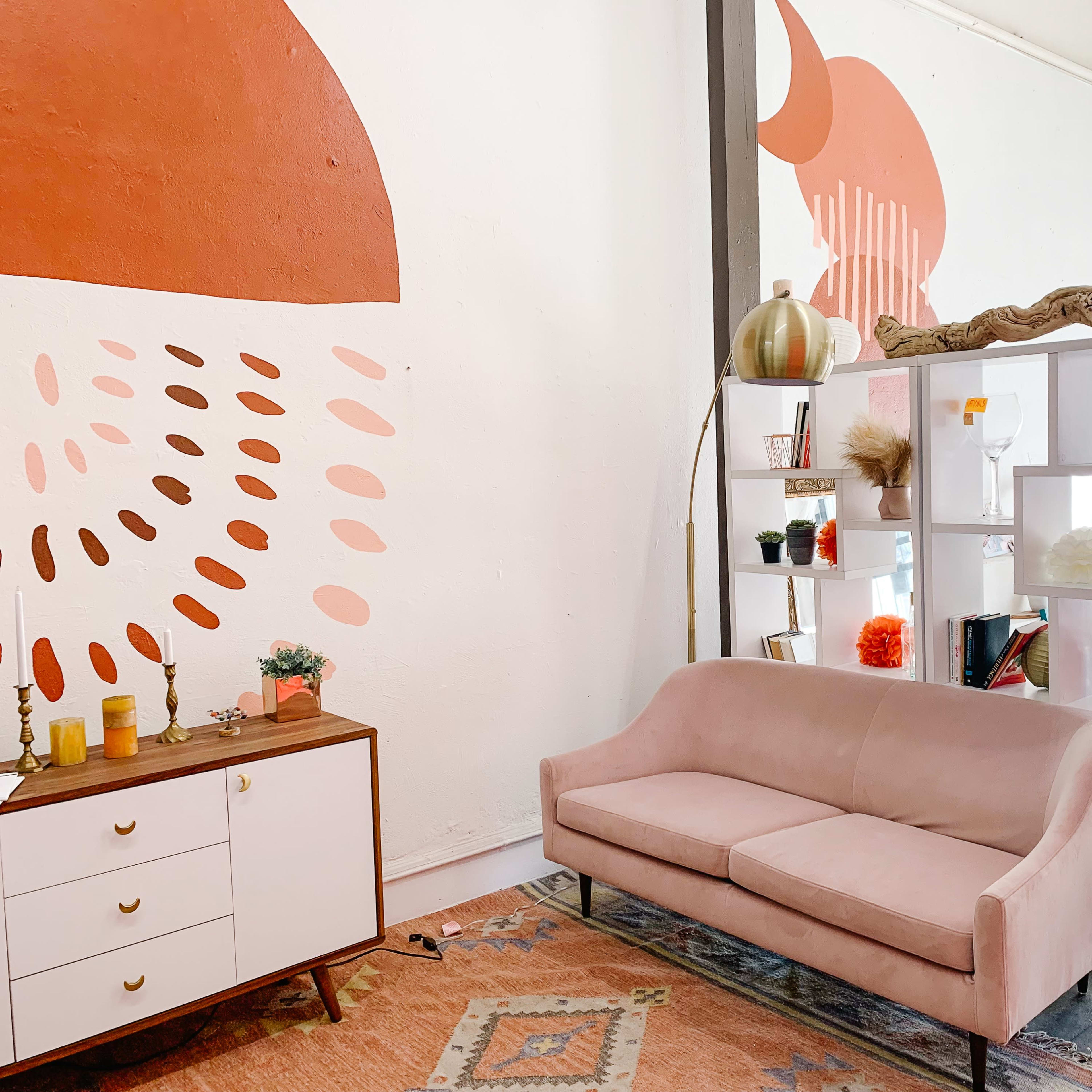 A modern interior featuring a pink sofa, a shelving unit with decorative items and books, and a wall painted with geometric patterns in warm tones.