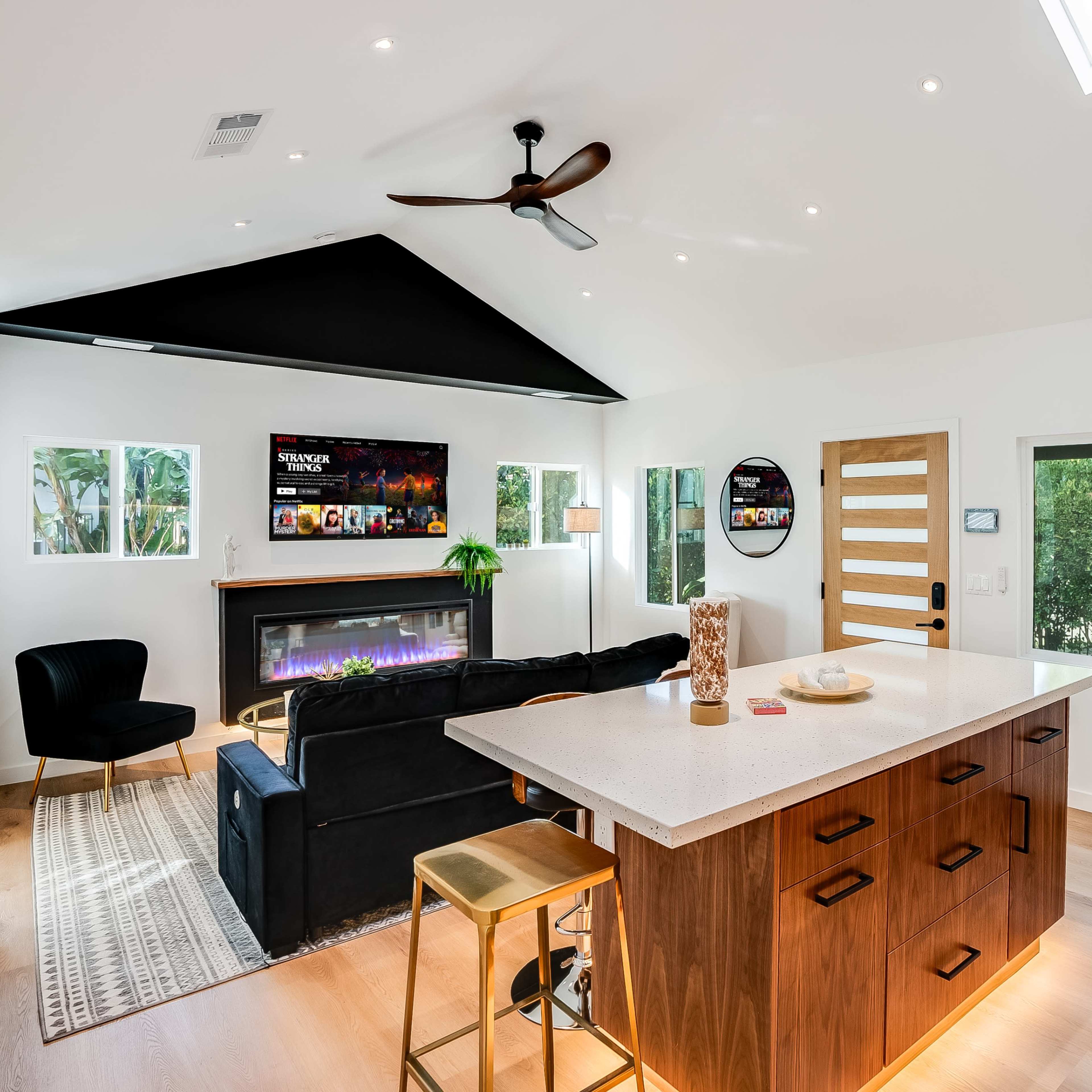 A modern open-concept living area features a kitchen island, sleek cabinetry, and a cozy seating arrangement with a fireplace and large windows.