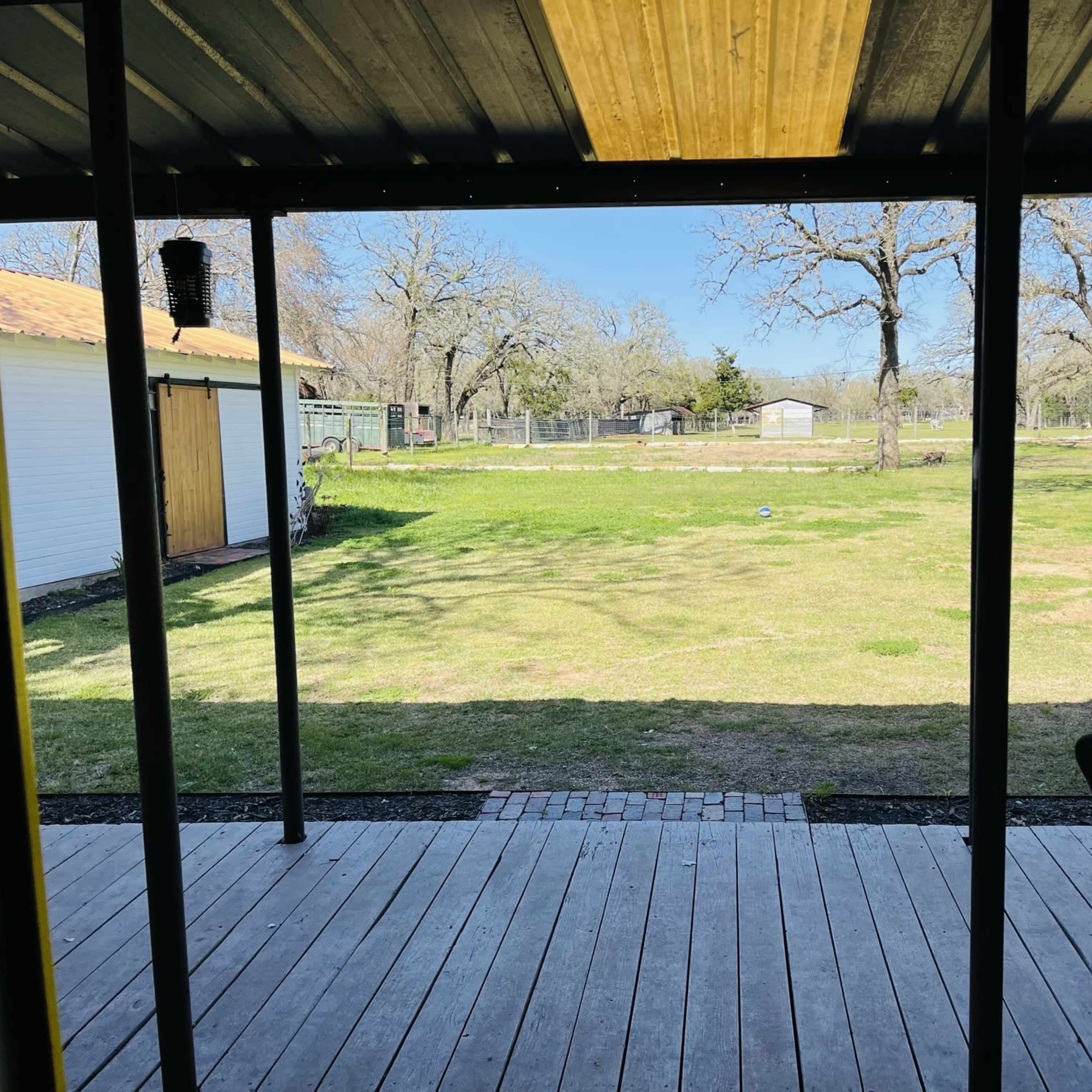 A view from a covered porch overlooking a grassy yard, with trees and a fenced area in the background.