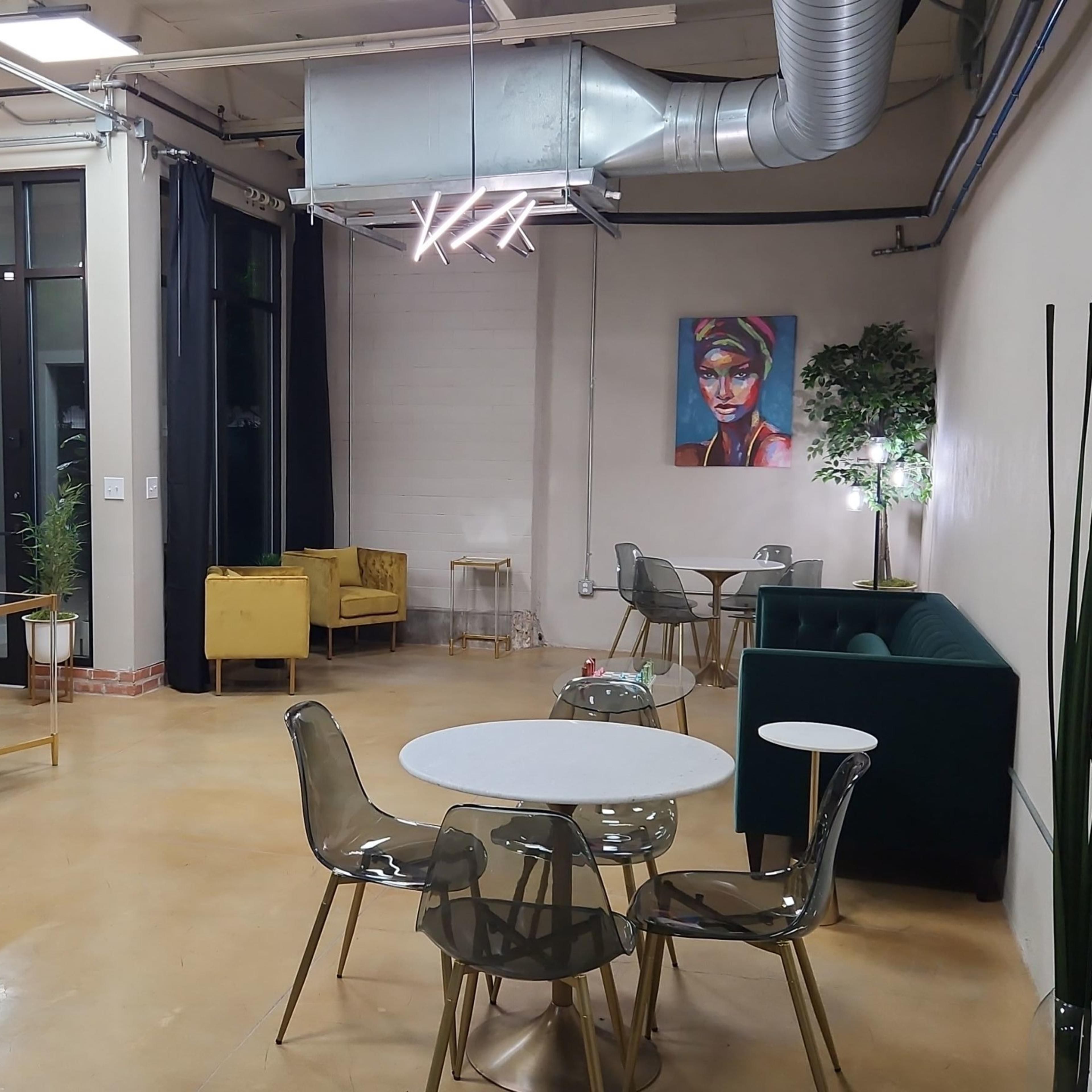 A modern, open interior space with a seating area that includes a green sofa, yellow chair, and several round tables surrounded by transparent chairs, along with art on the wall and plants.