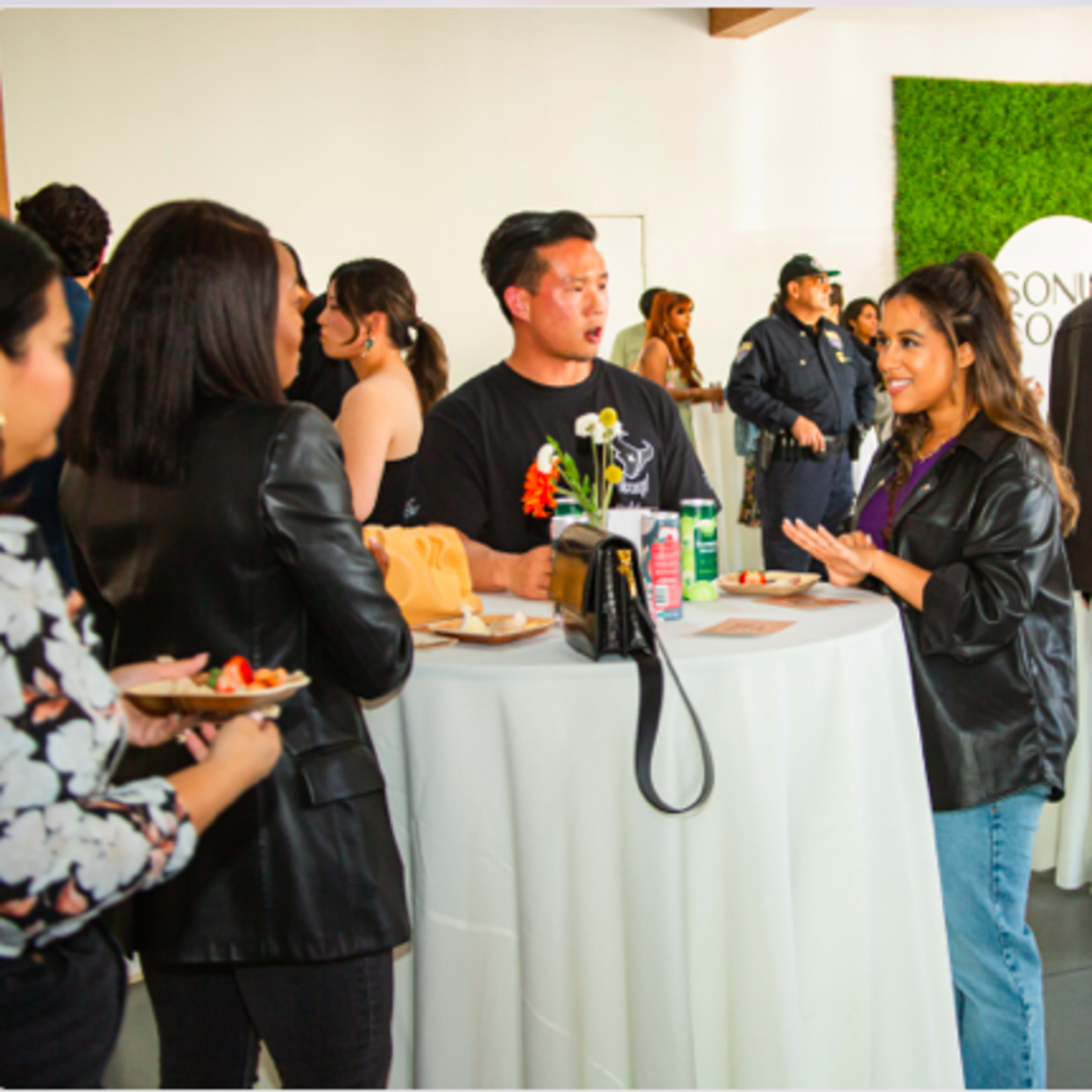 A social gathering with guests engaging in conversation and mingling around food and drink tables in a well-lit venue.