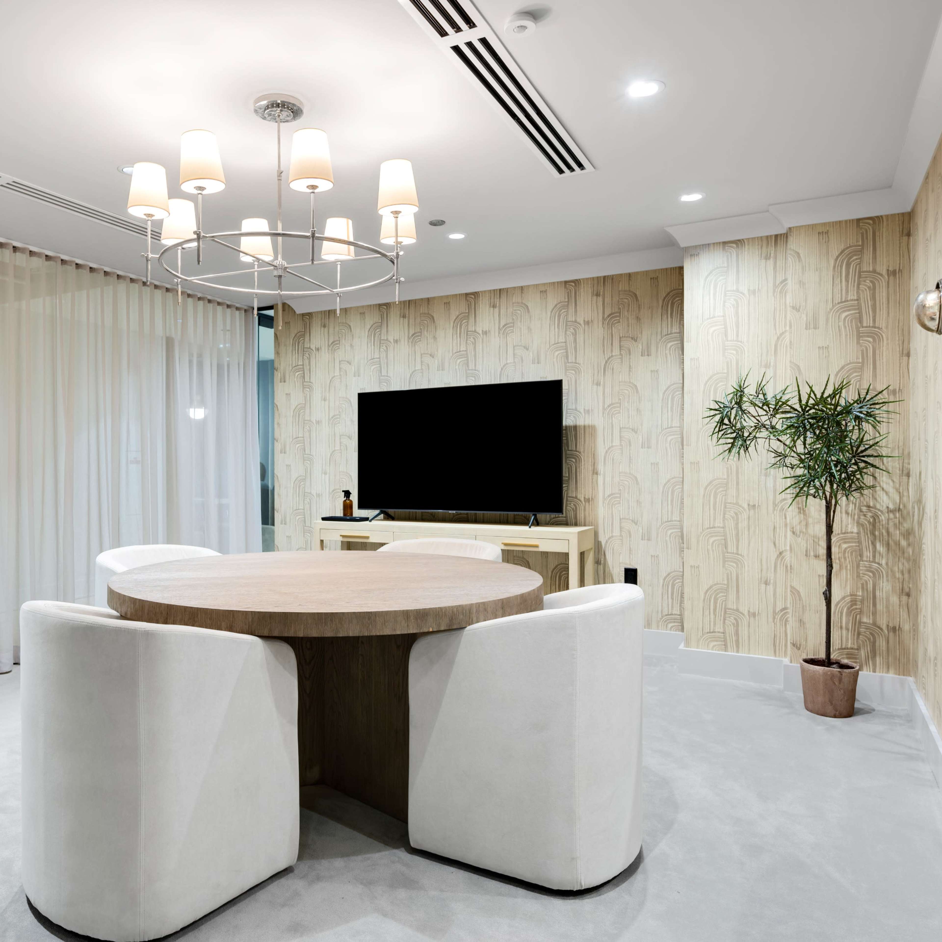 A modern dining area featuring a round wooden table surrounded by four upholstered chairs, a large flat-screen TV on a light-colored cabinet, and a potted plant in the corner.