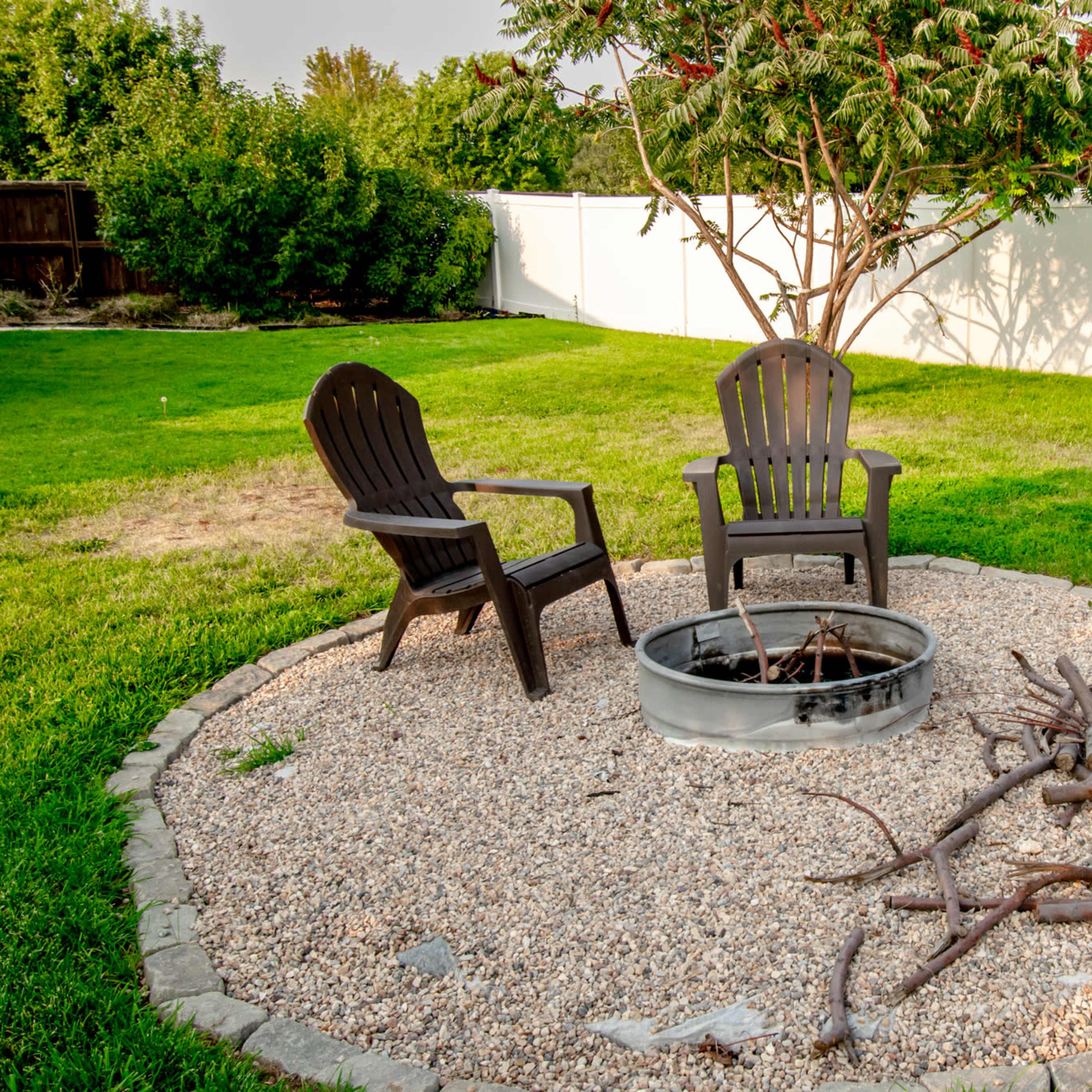 A circular gravel patio features two black Adirondack chairs around a fire pit, with a pile of sticks nearby, set against a backdrop of a grassy yard and a white fence.
