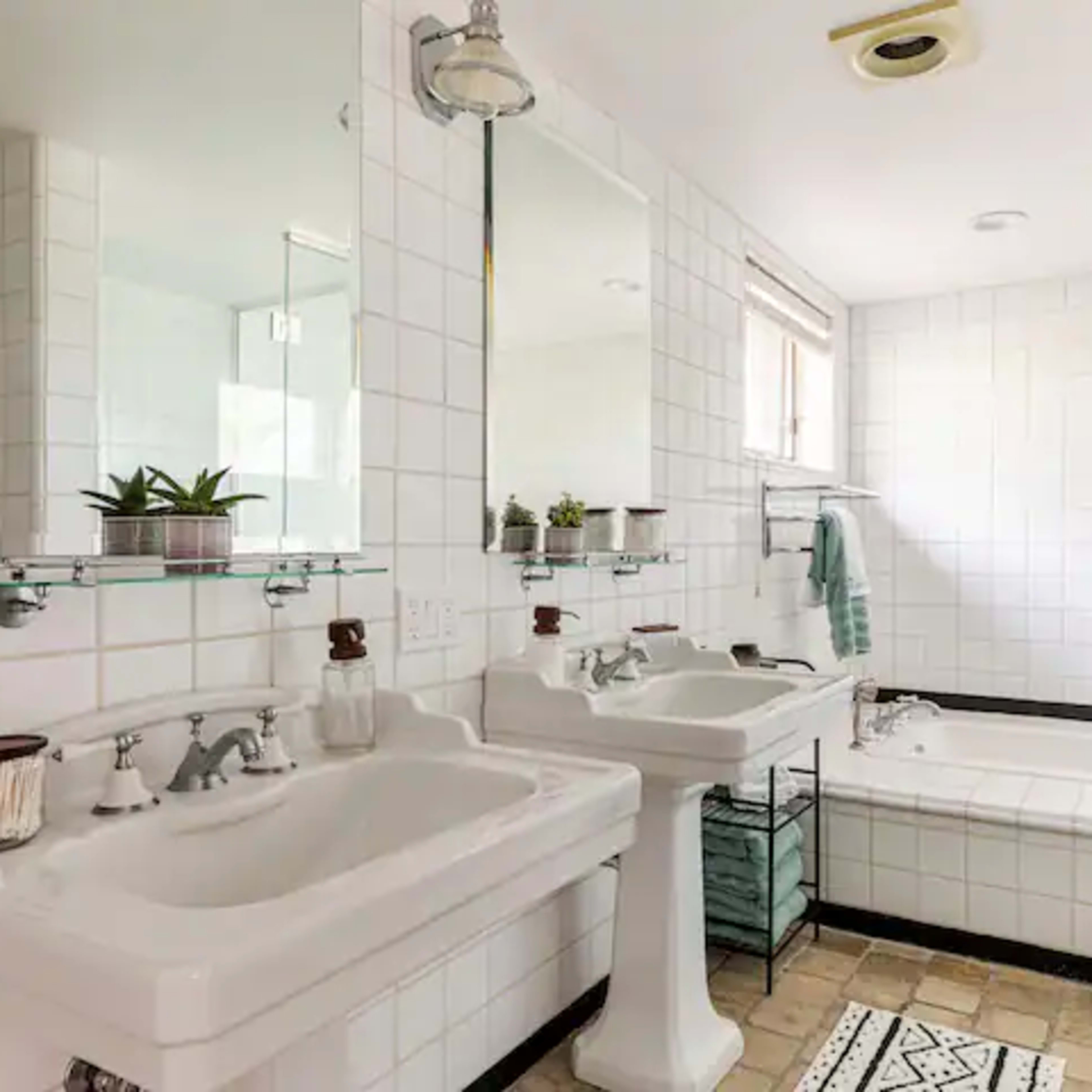 A bright bathroom featuring two pedestal sinks, a bathtub, and white tiled walls with black accents.