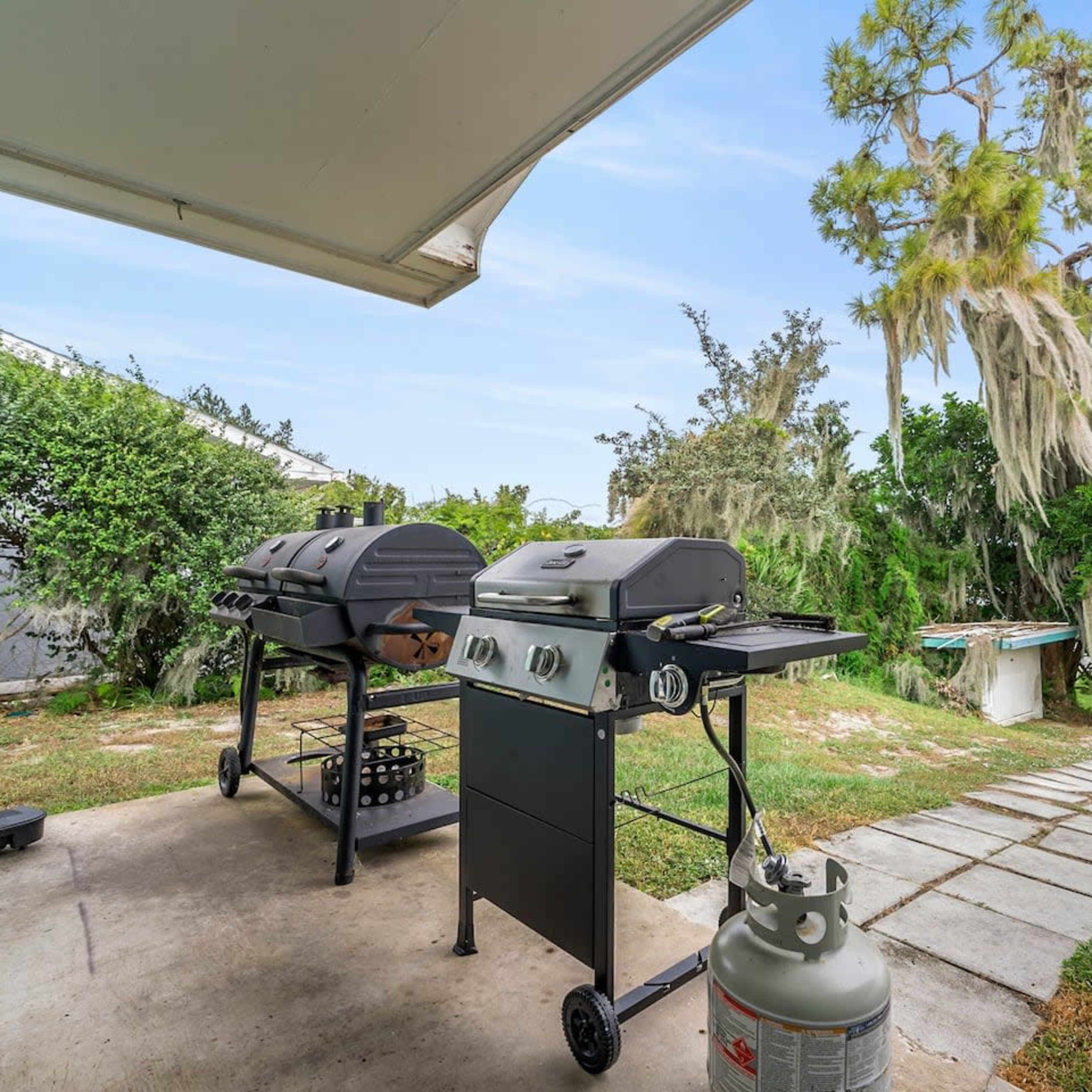 Two gas grills are set up on a concrete patio, overlooking a body of water with trees in the background.