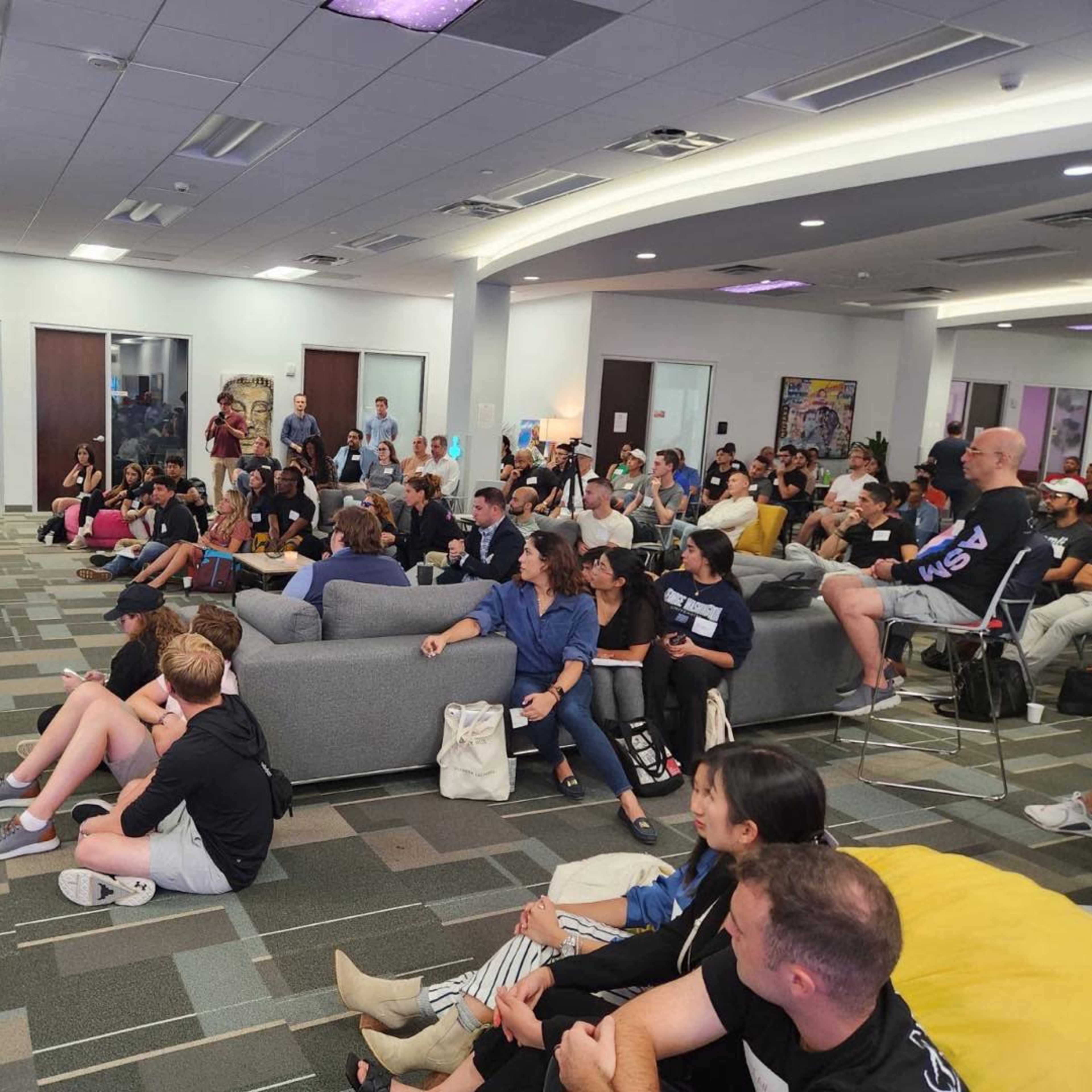 A large group of people listens to a speaker in a modern office space with gray sofas and colorful bean bags.