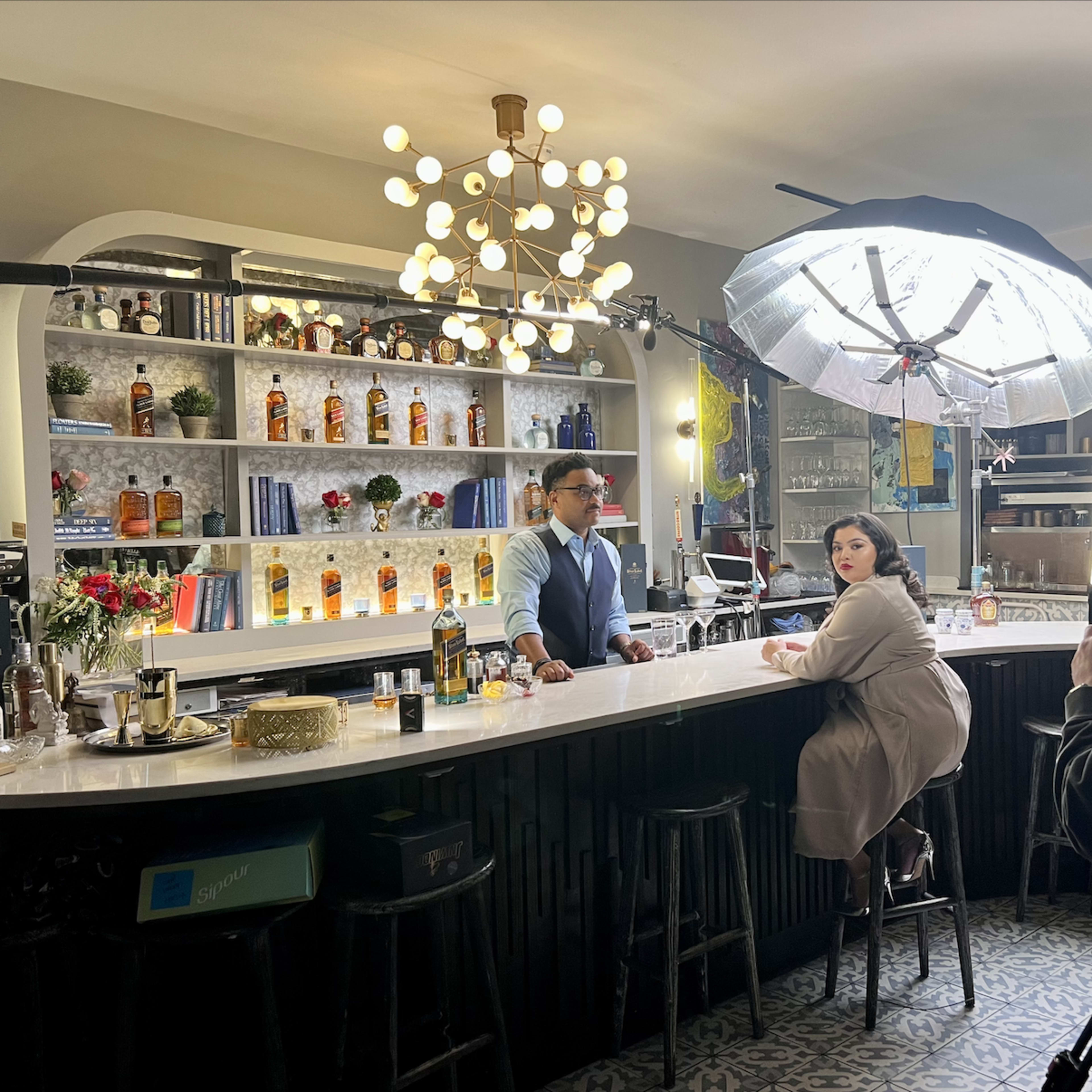 A bartender and a woman sit at a well-lit bar surrounded by shelves of various spirits and decorations, with a large softbox light directing focus on them.