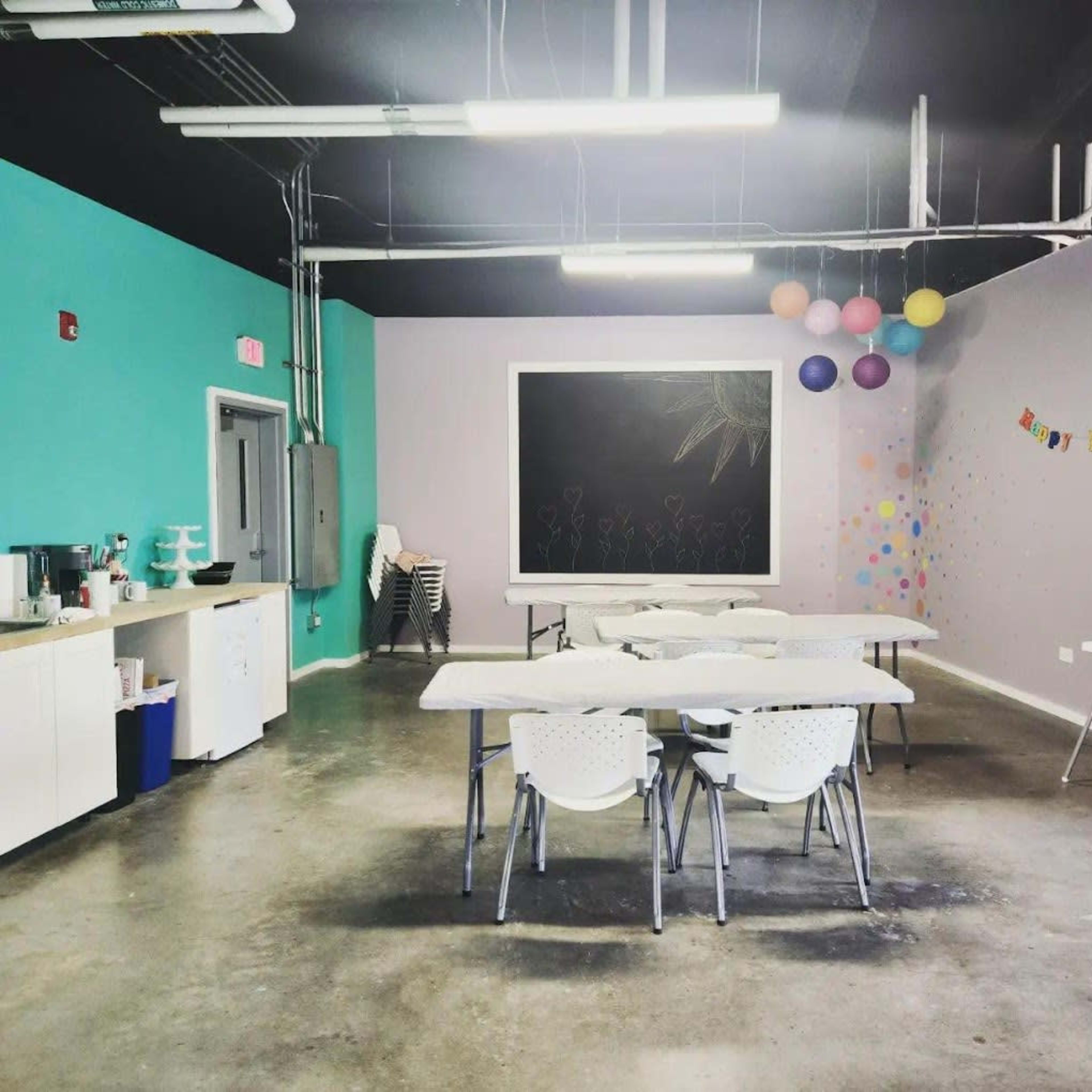 A spacious room with multiple tables and chairs arranged, a chalkboard on the wall, and colorful decorations including balloons and a birthday banner.