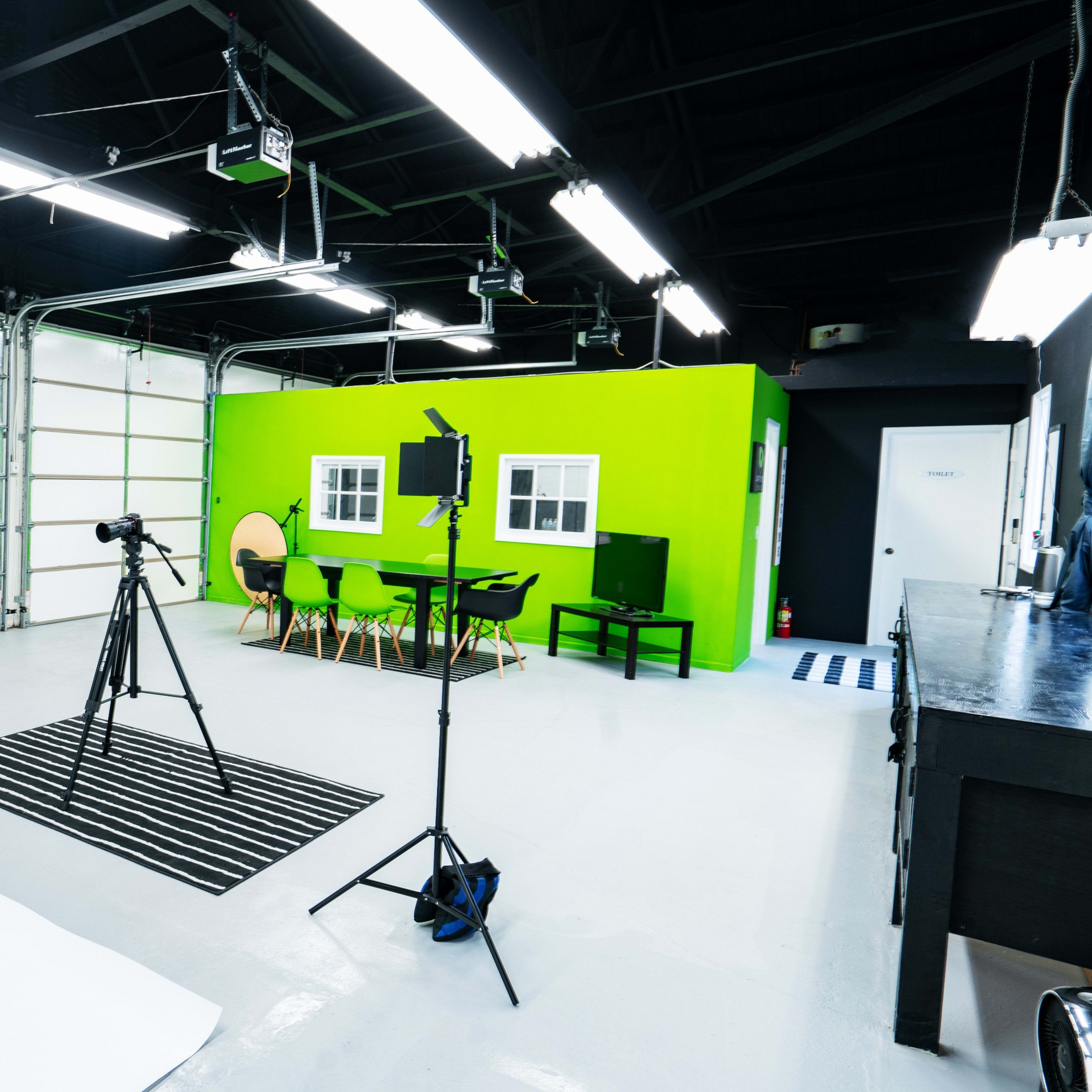 A photography studio with a green wall, various lighting equipment, a white backdrop, a black table, and a green chair arranged in the space.