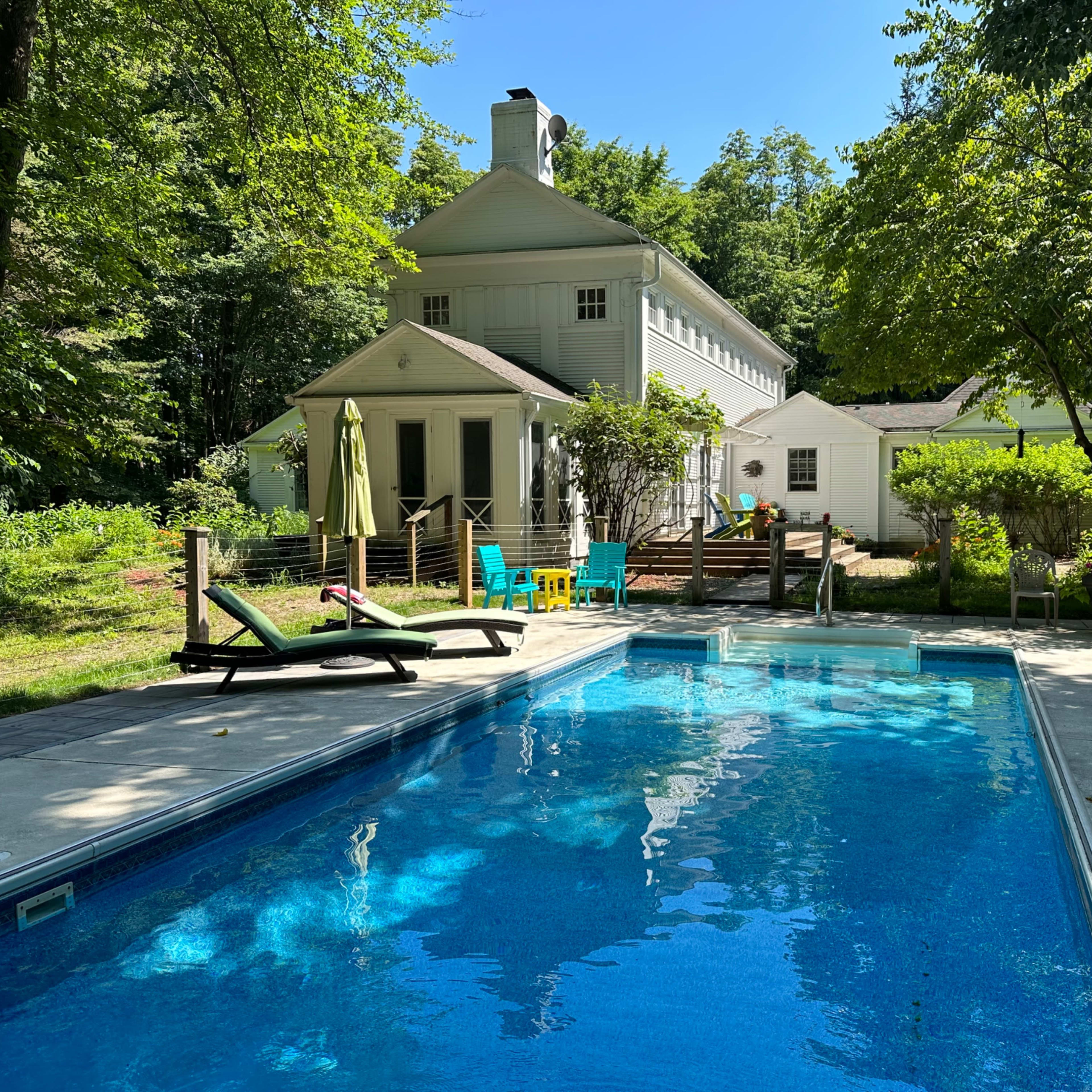 A clear blue swimming pool is situated in the foreground, with a white house and green trees in the background.