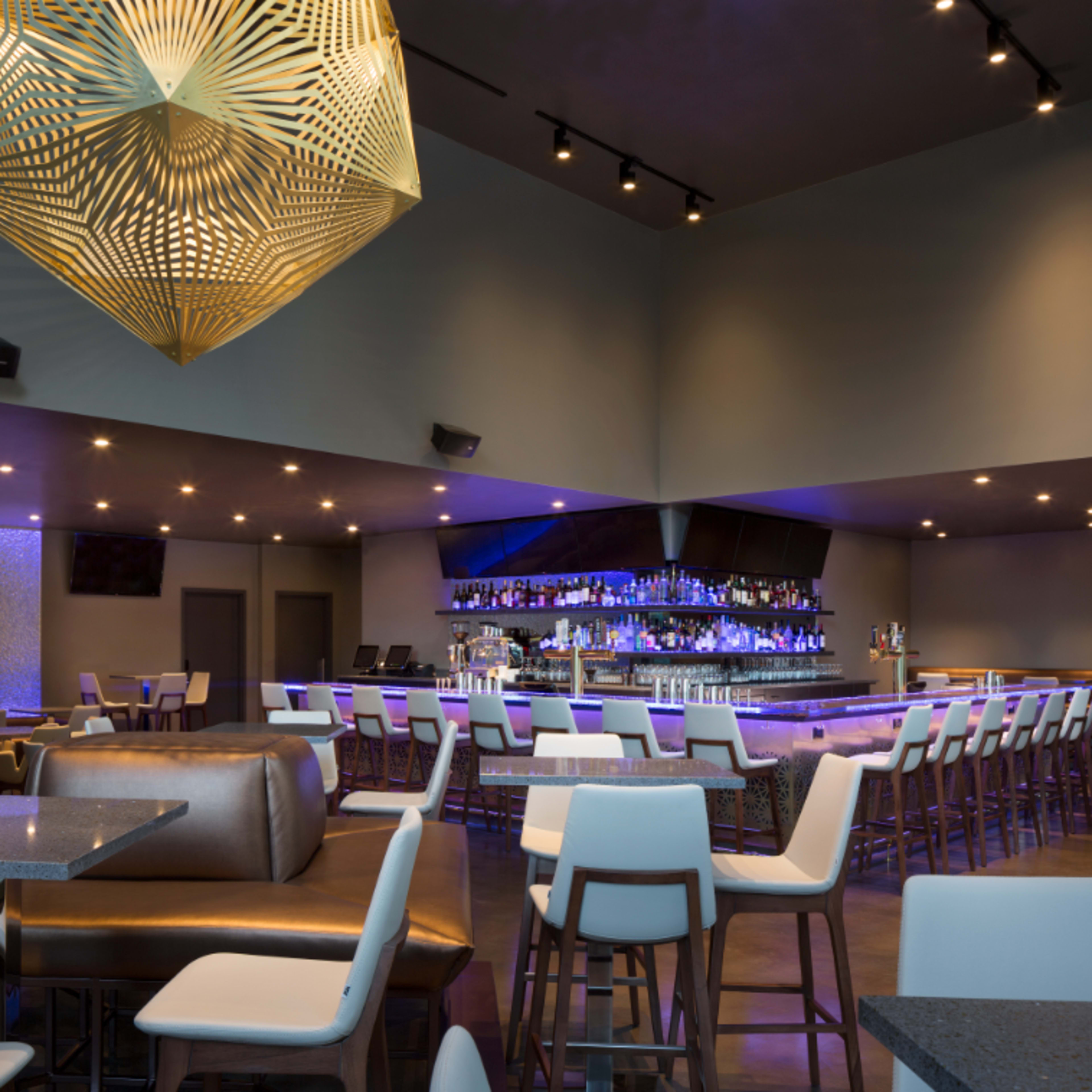 A modern bar and dining area with a stylish geometric light fixture overhead, a long bar stocked with drinks, and multiple seating arrangements.