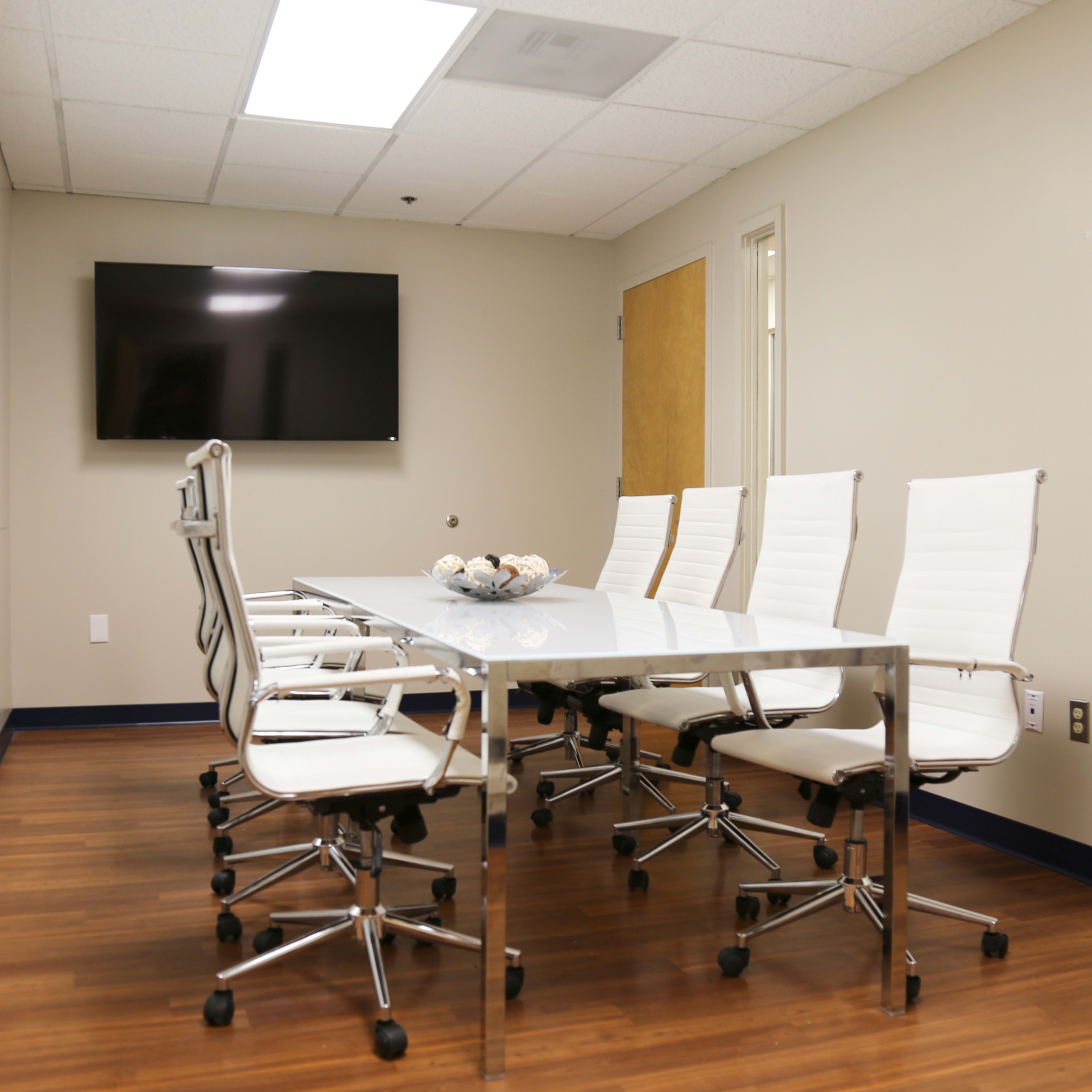 A modern conference room with a glass table and white rolling chairs, featuring a wall-mounted TV and a light-colored wall.