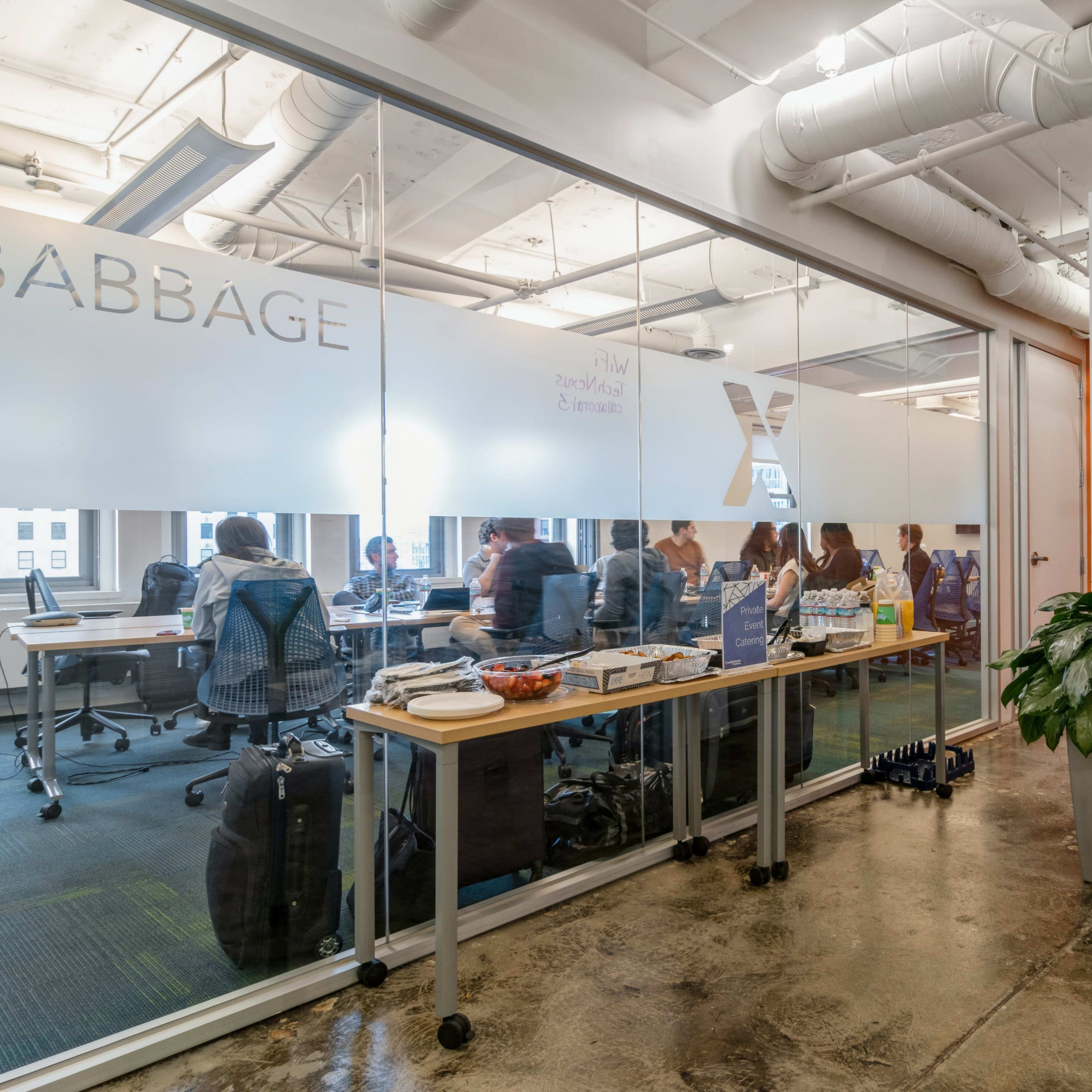 A conference room is visible through glass walls, with a group of people seated at a long table engaged in a meeting, while a plant and a side table with various items are positioned in the foreground.