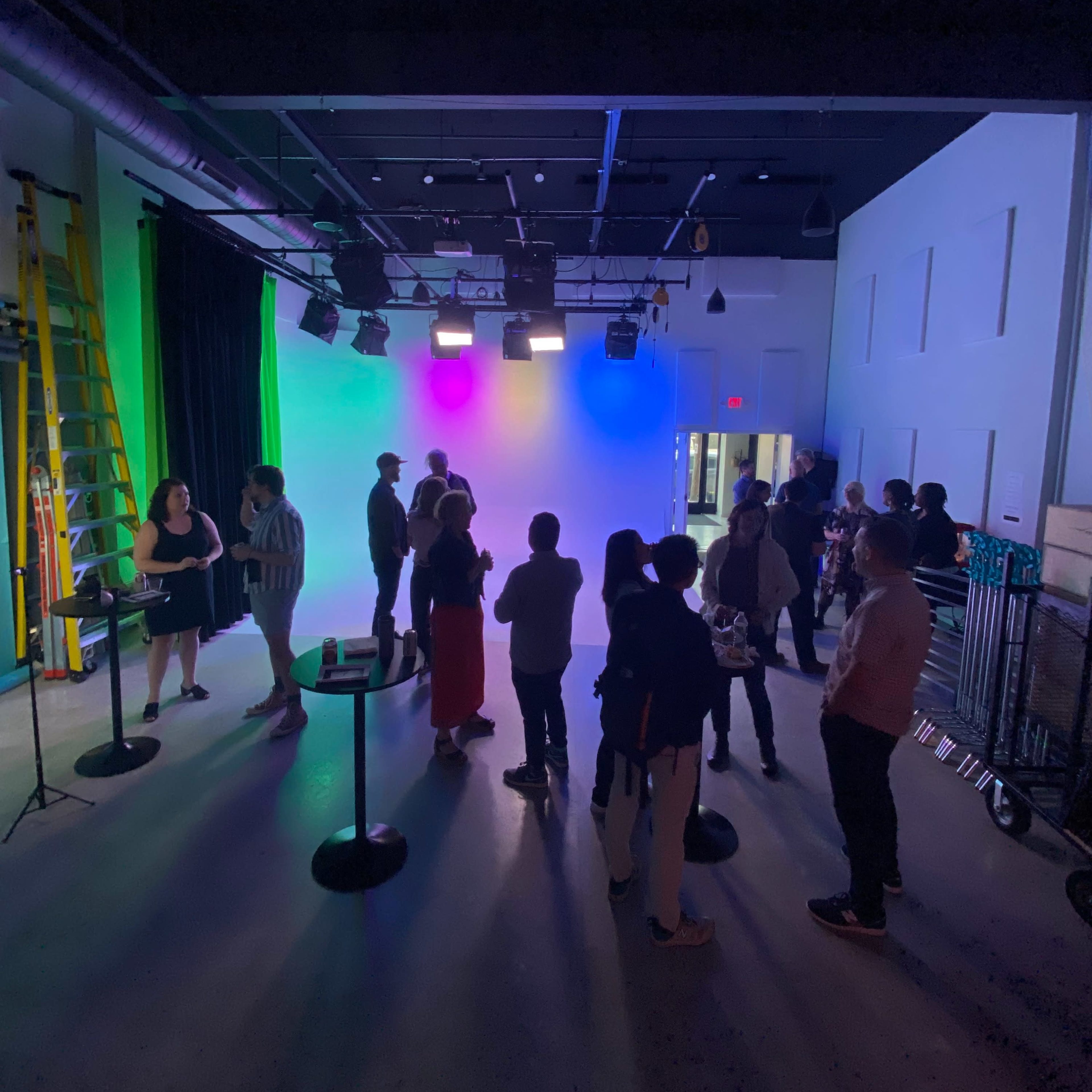 A group of people gathers in a brightly lit indoor space with colored backdrop lighting and various decorations.