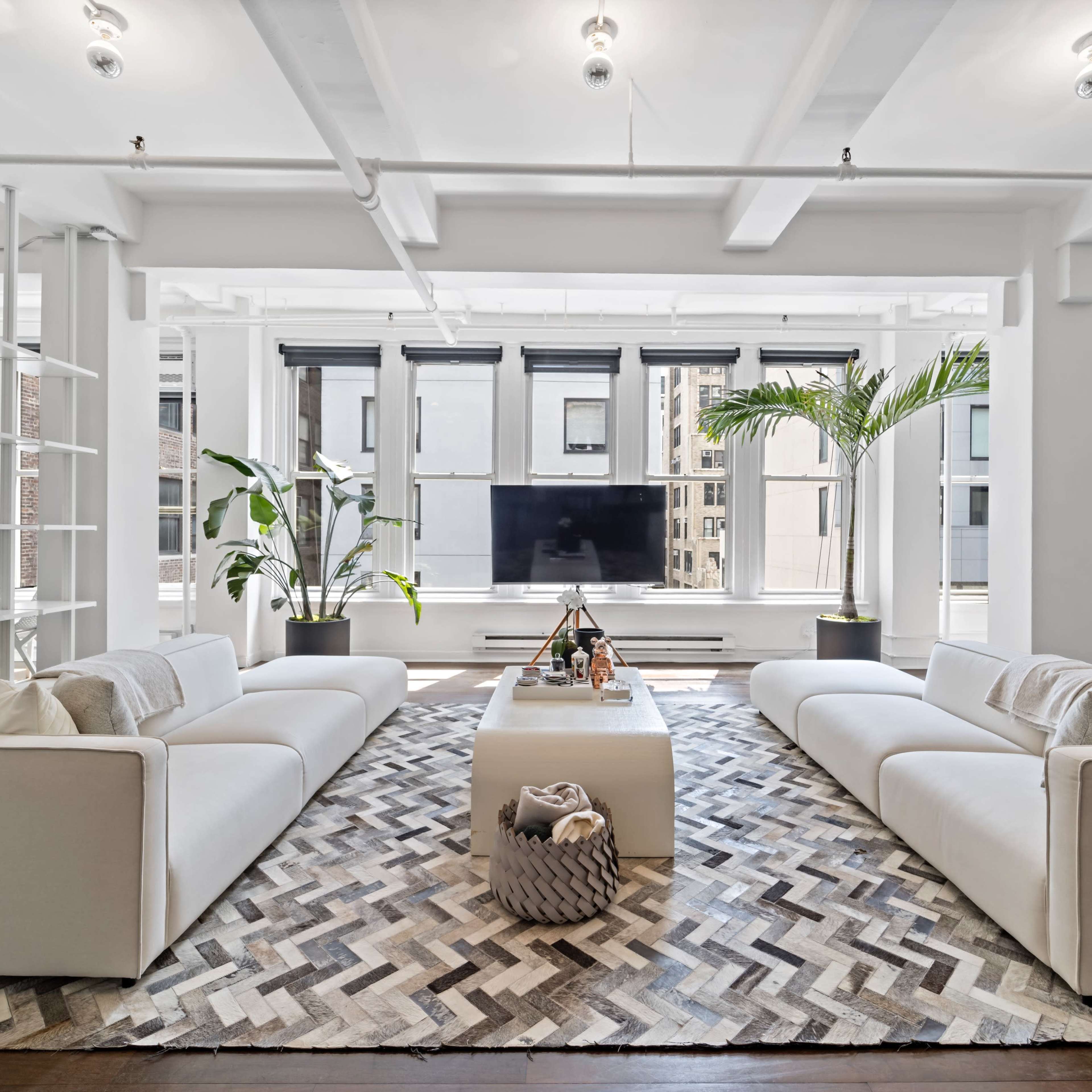 A spacious living area features two white sofas facing a central coffee table, with large windows providing natural light and greenery from potted plants.