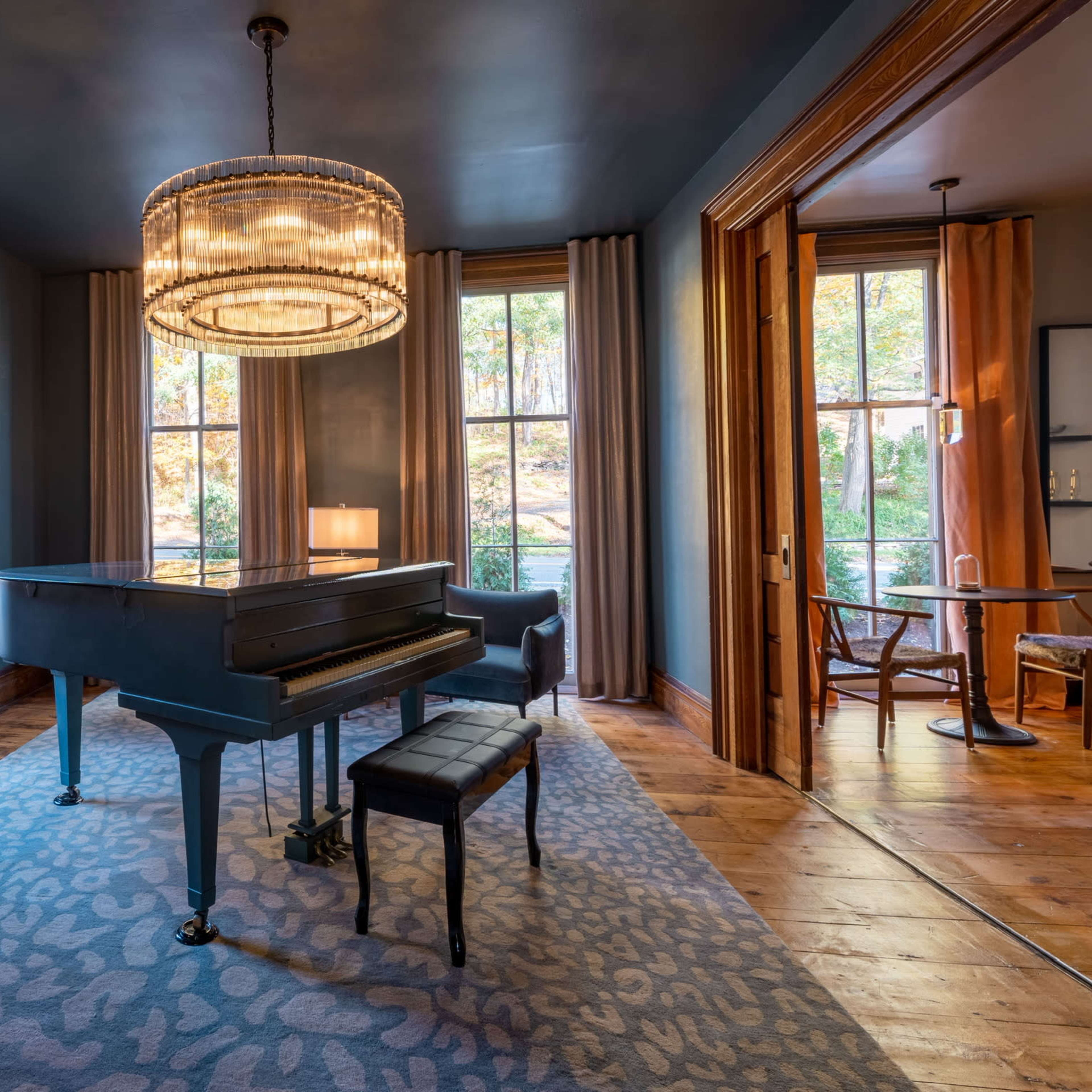 A grand piano sits in a room with dark walls, large windows, and a chandelier, surrounded by elegant furnishings and a patterned rug.