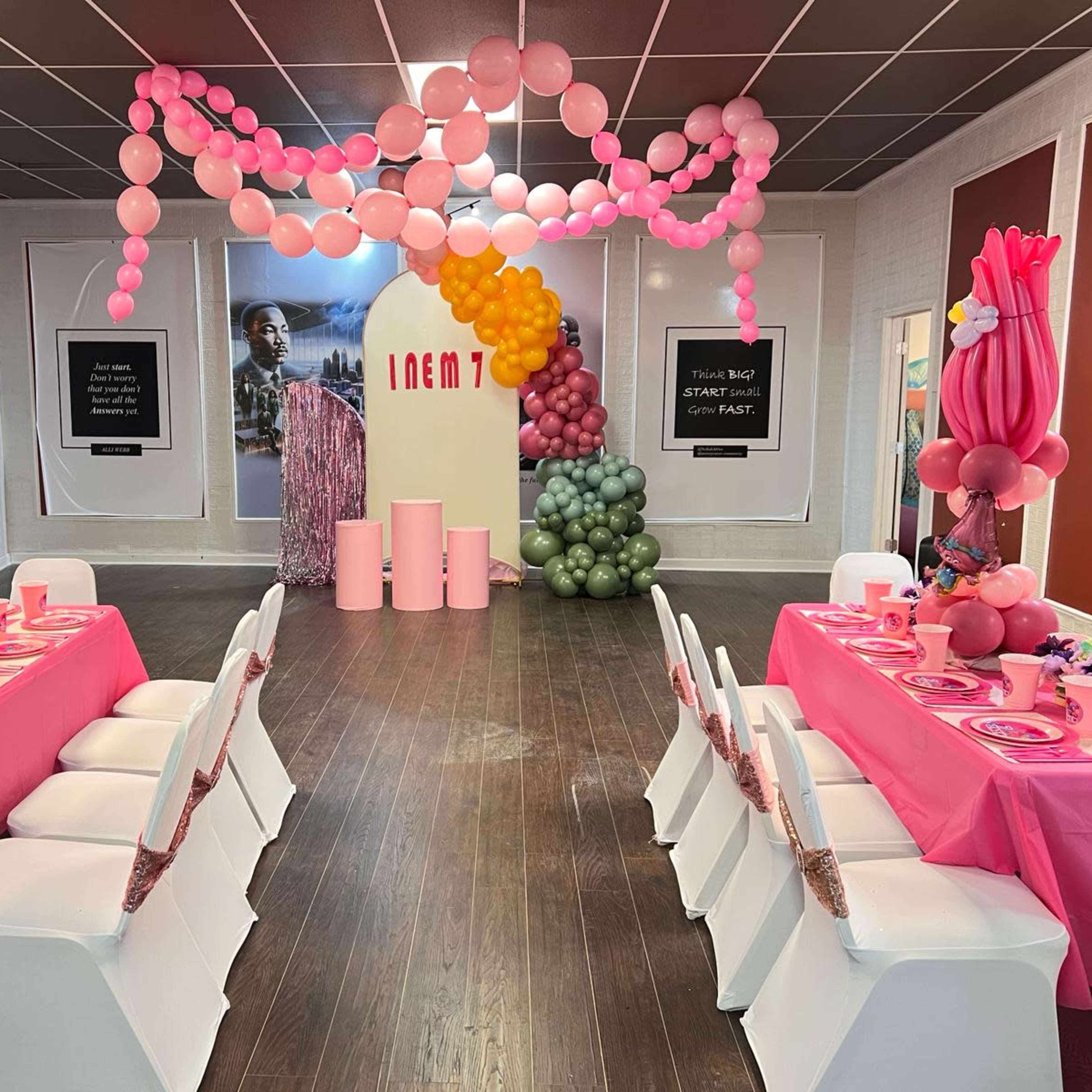 A decorated event space featuring tables set with pink tablecloths and colorful centerpieces, adorned with balloon arches and themed decorations.
