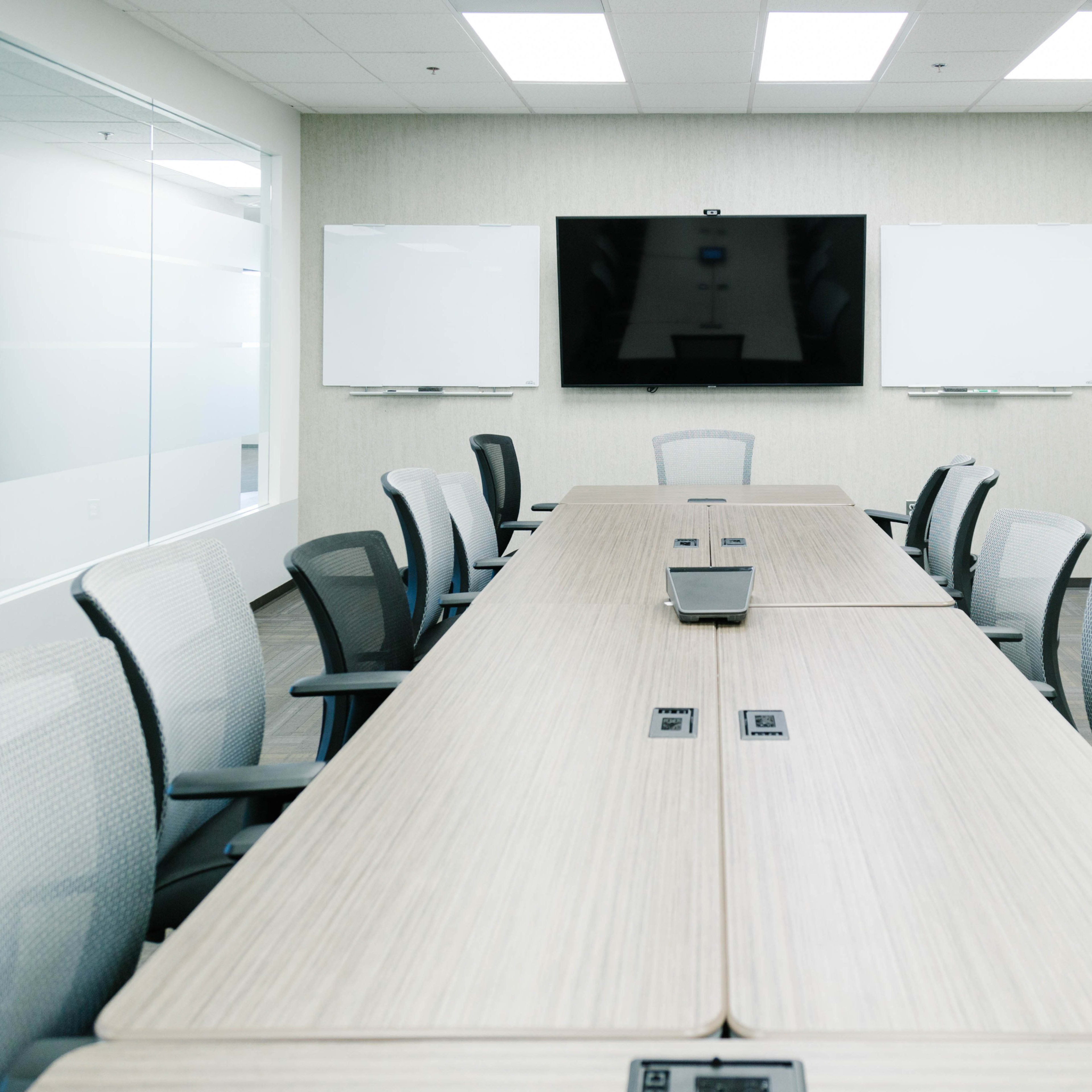A modern conference room features a long wooden table, multiple office chairs, and a large wall-mounted screen with whiteboards on either side.