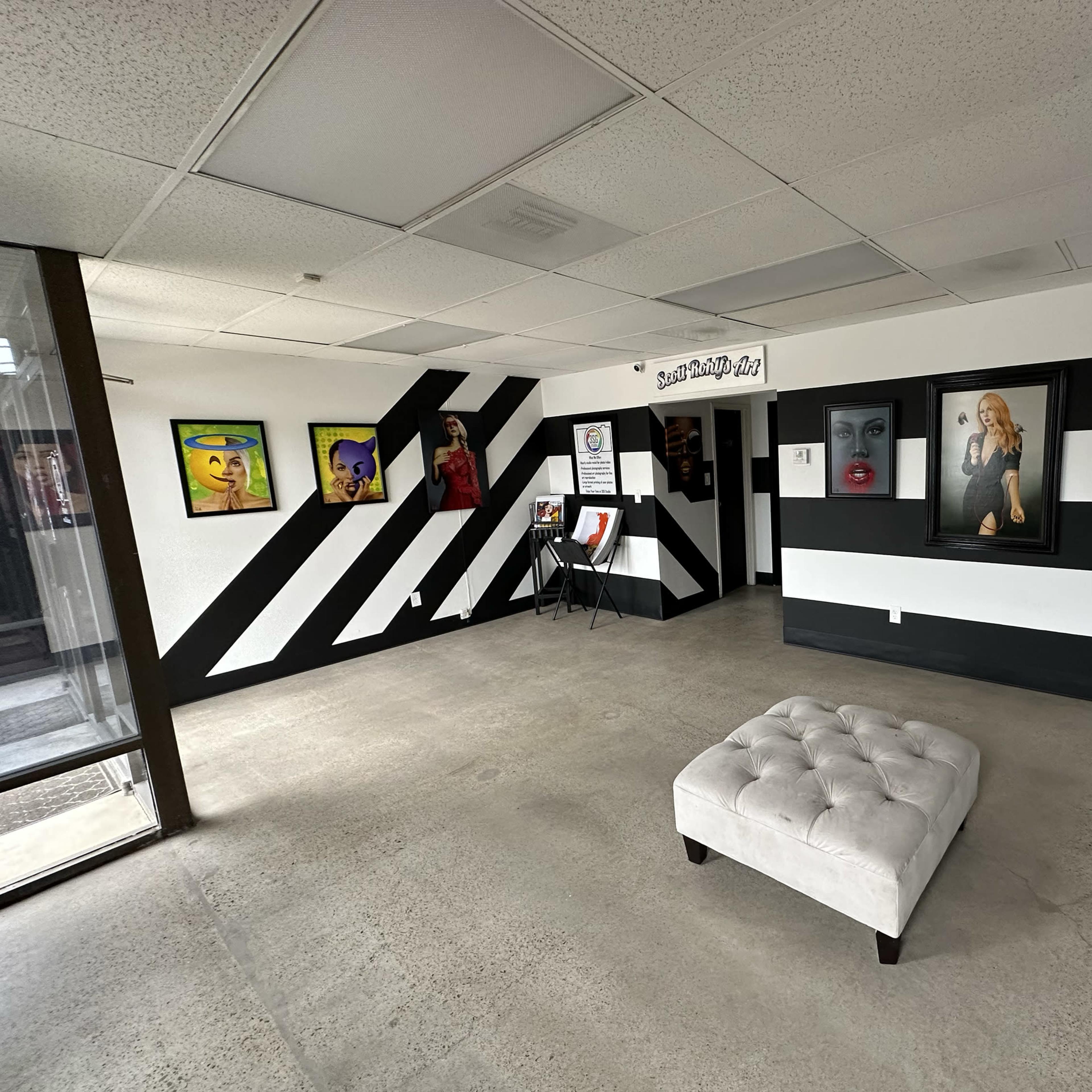 An art gallery with black and white striped walls, featuring various framed artworks on display and a tufted ottoman in the center.