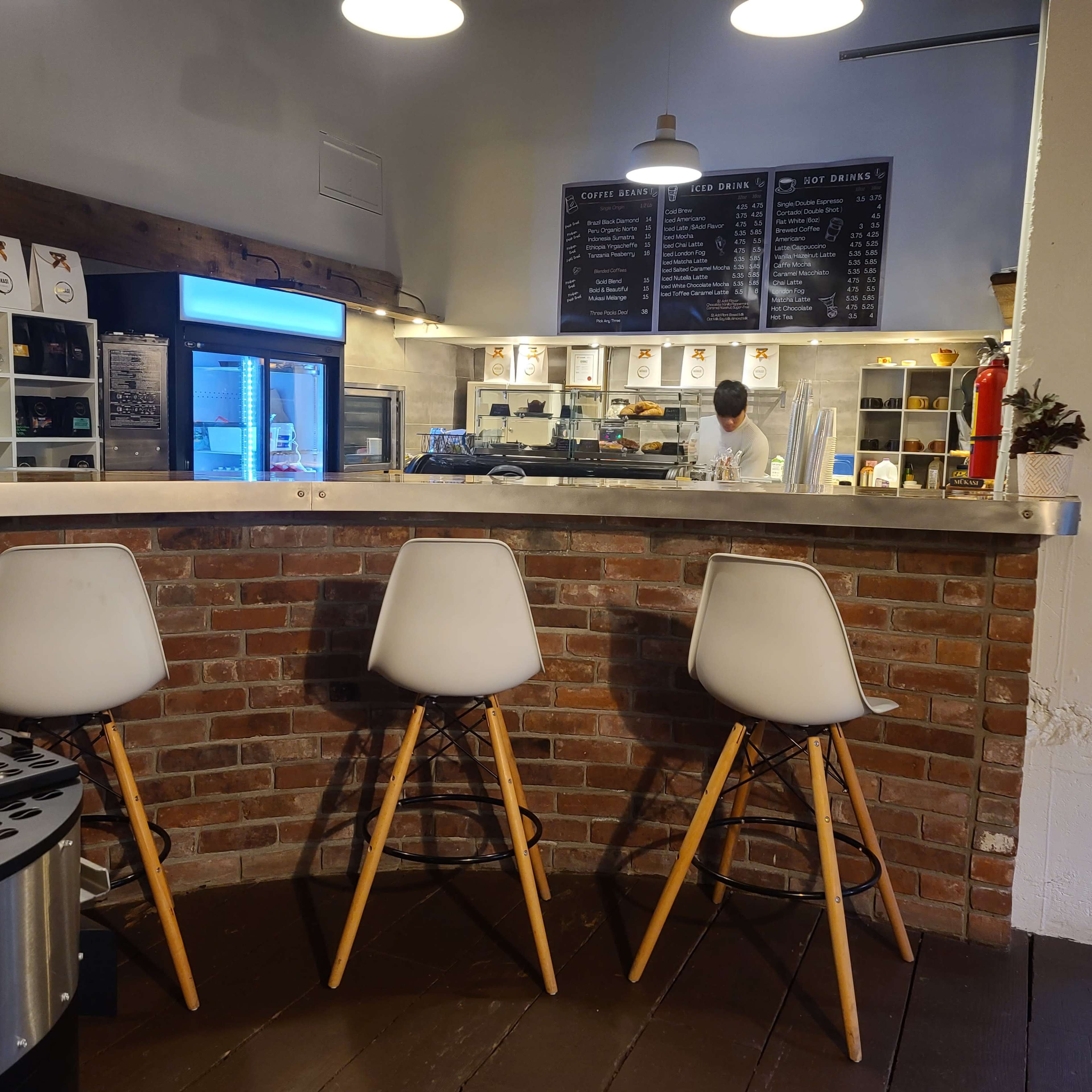 A bar area with three white chairs sits in front of a kitchen space featuring a chef and a menu board on the wall.