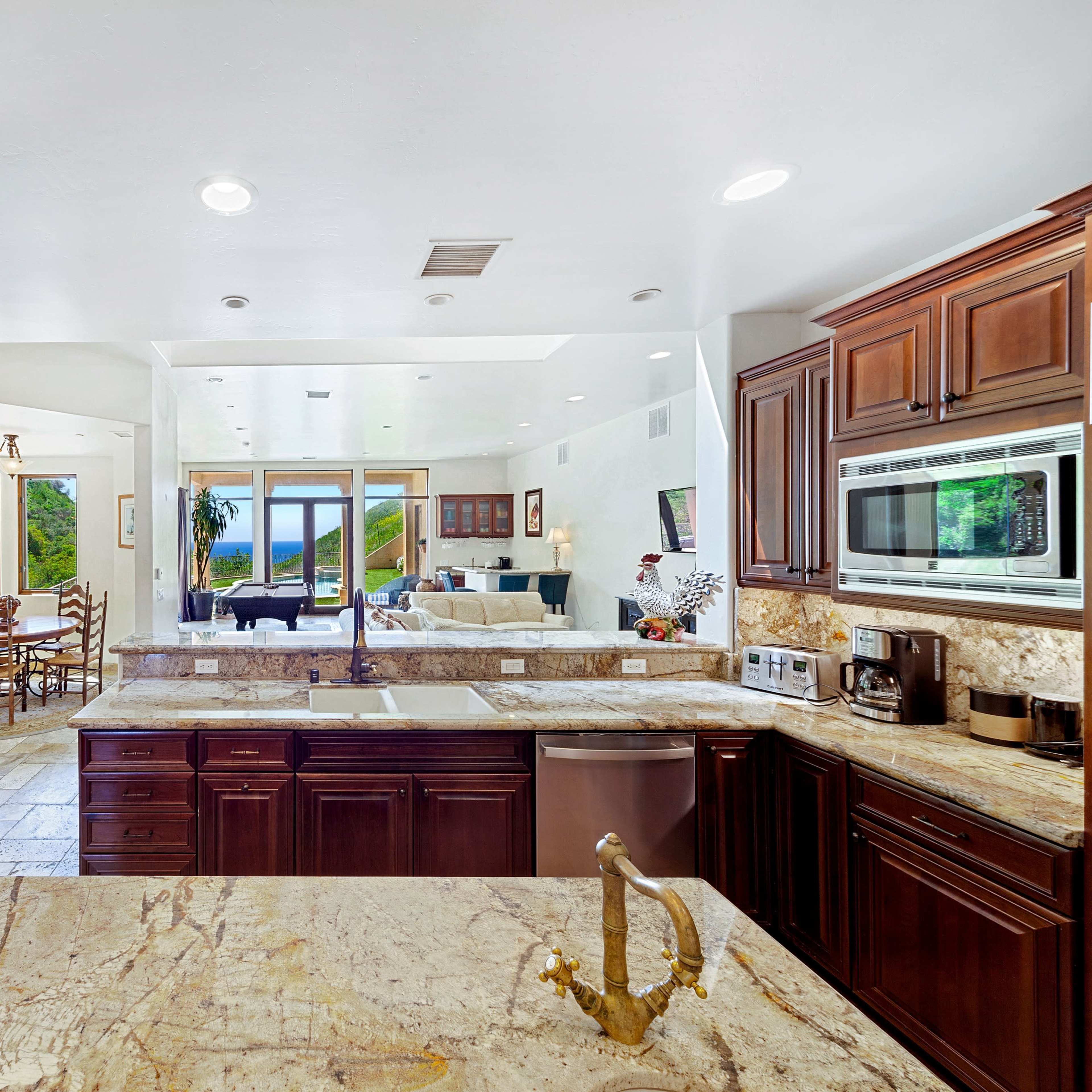 A spacious kitchen features wooden cabinetry, a marble countertop, and opens to a living area with views of the outdoors.
