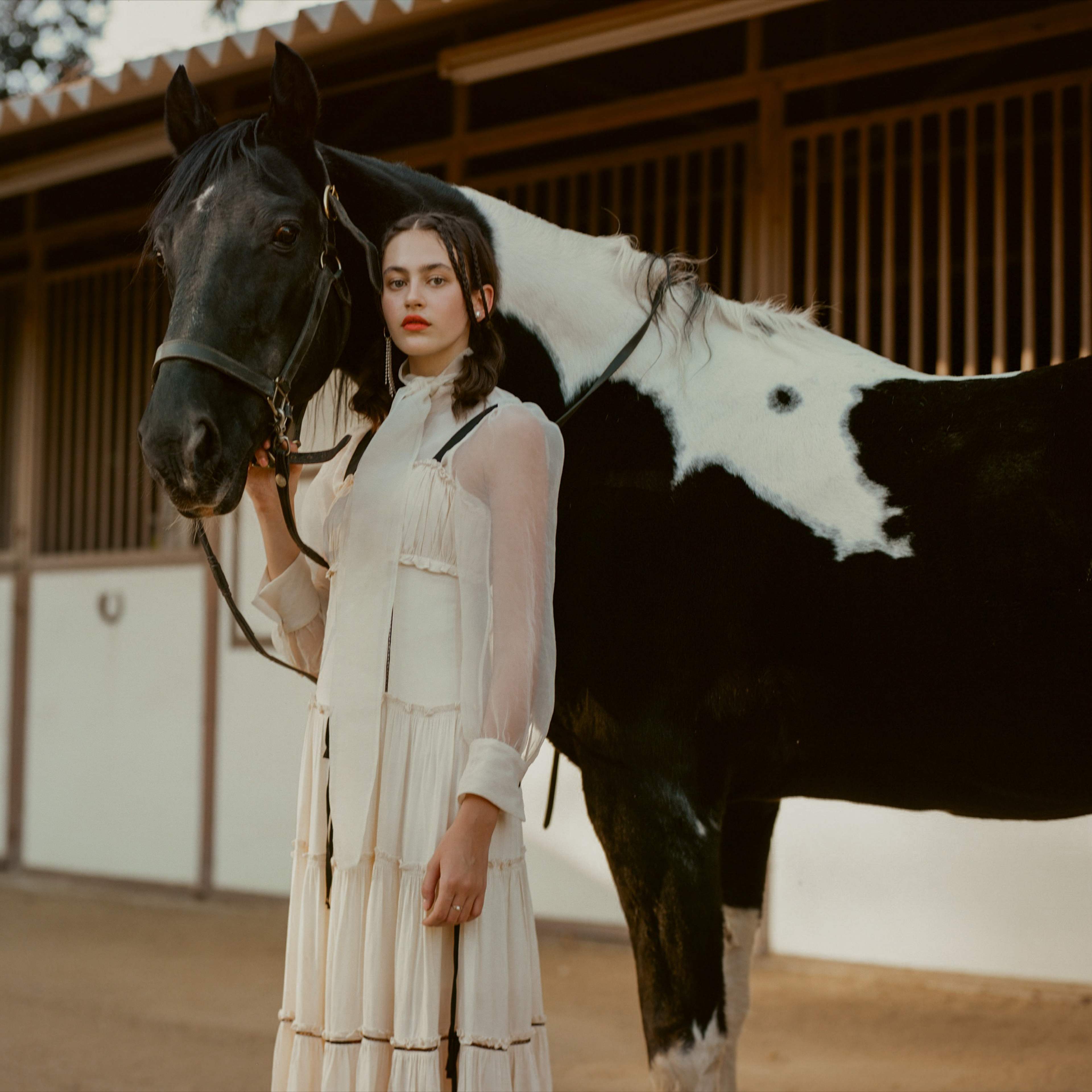 A woman in a white dress stands next to a black and white horse in a barn setting.
