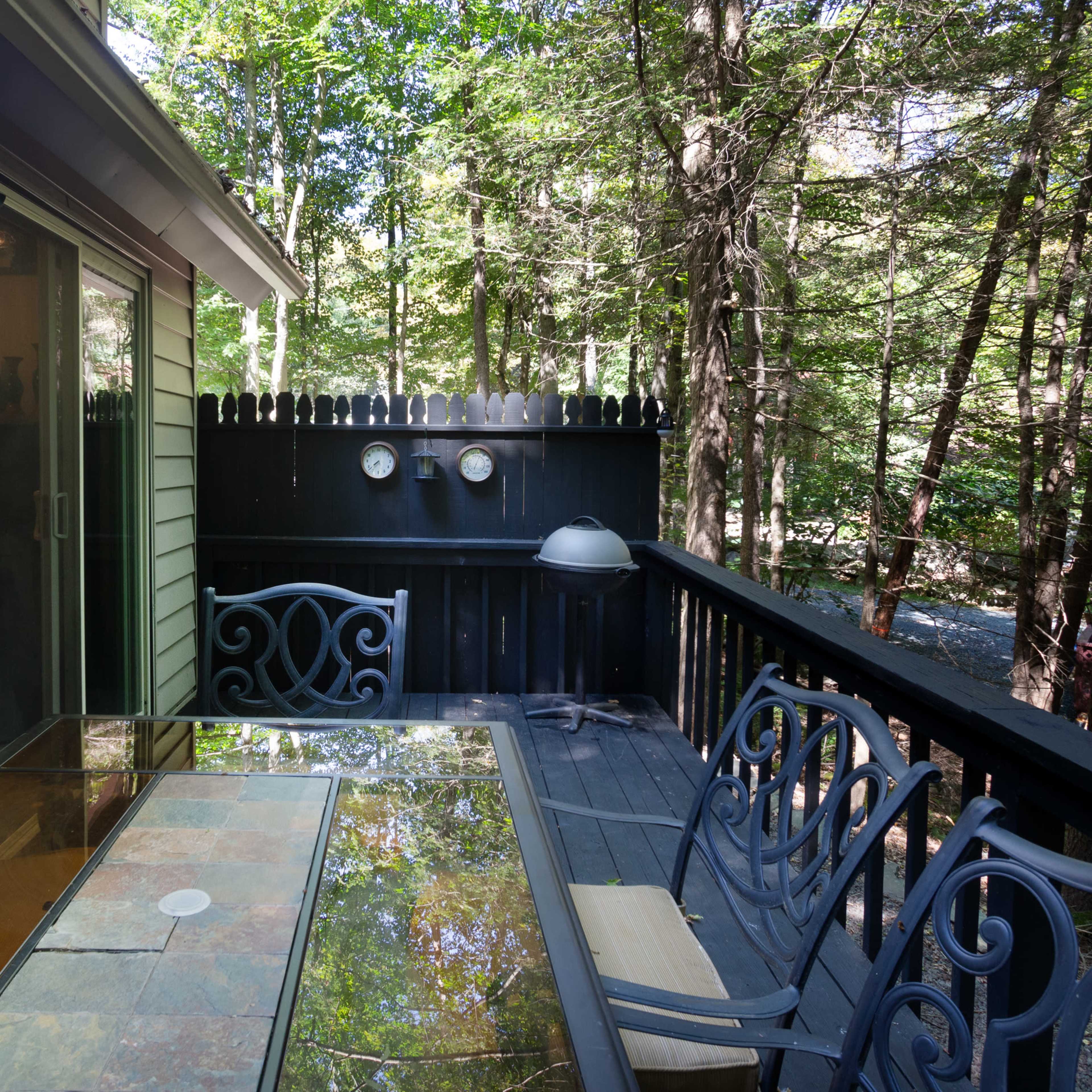 A wooden deck features a glass-topped table surrounded by metal chairs, with a view of trees and a black fence in the background.