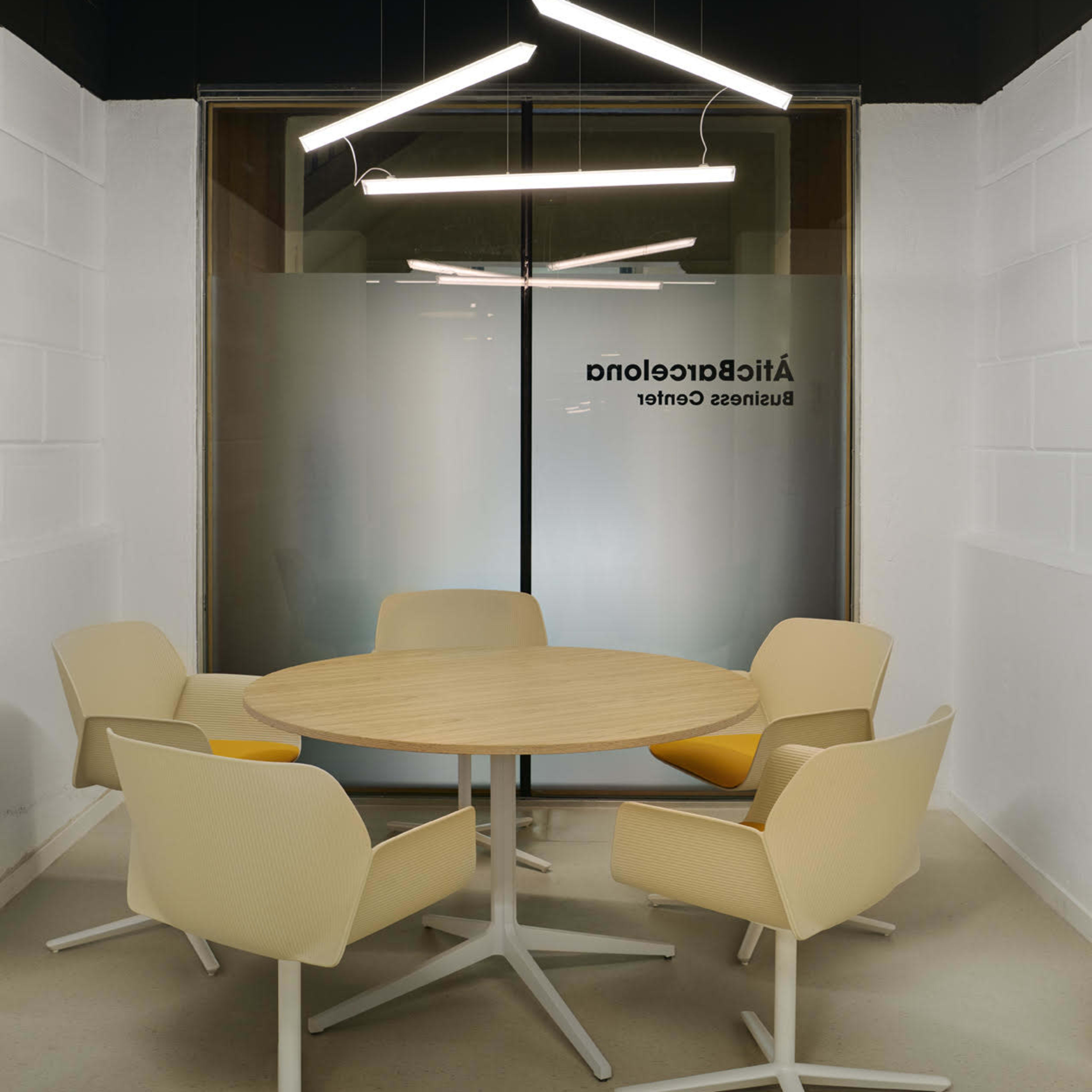 A round wooden table surrounded by four beige chairs is set in a modern conference room with a glass wall.