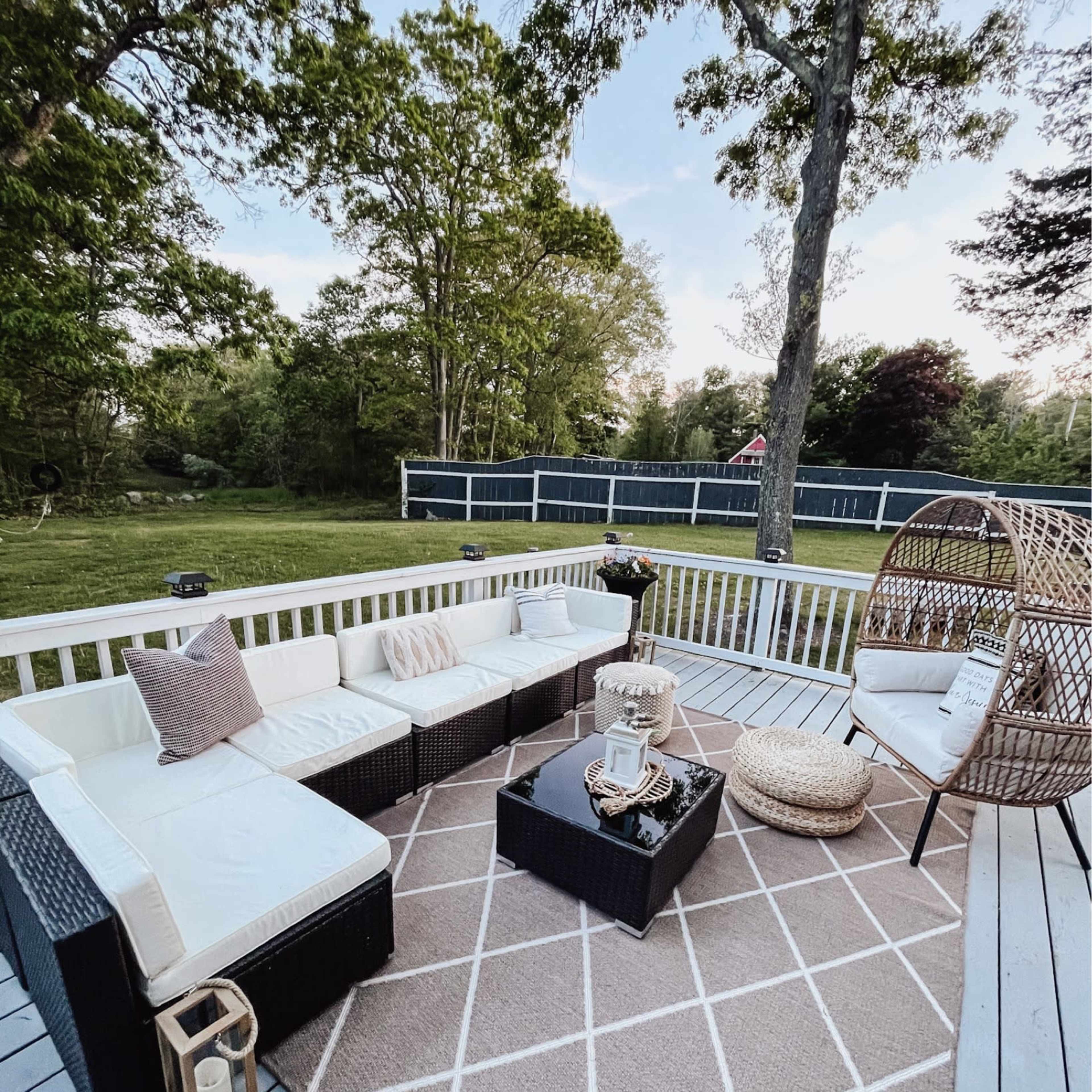 A patio area featuring a sectional couch, a round chair, a coffee table on a geometric-patterned rug, and surrounded by trees and a grassy yard.
