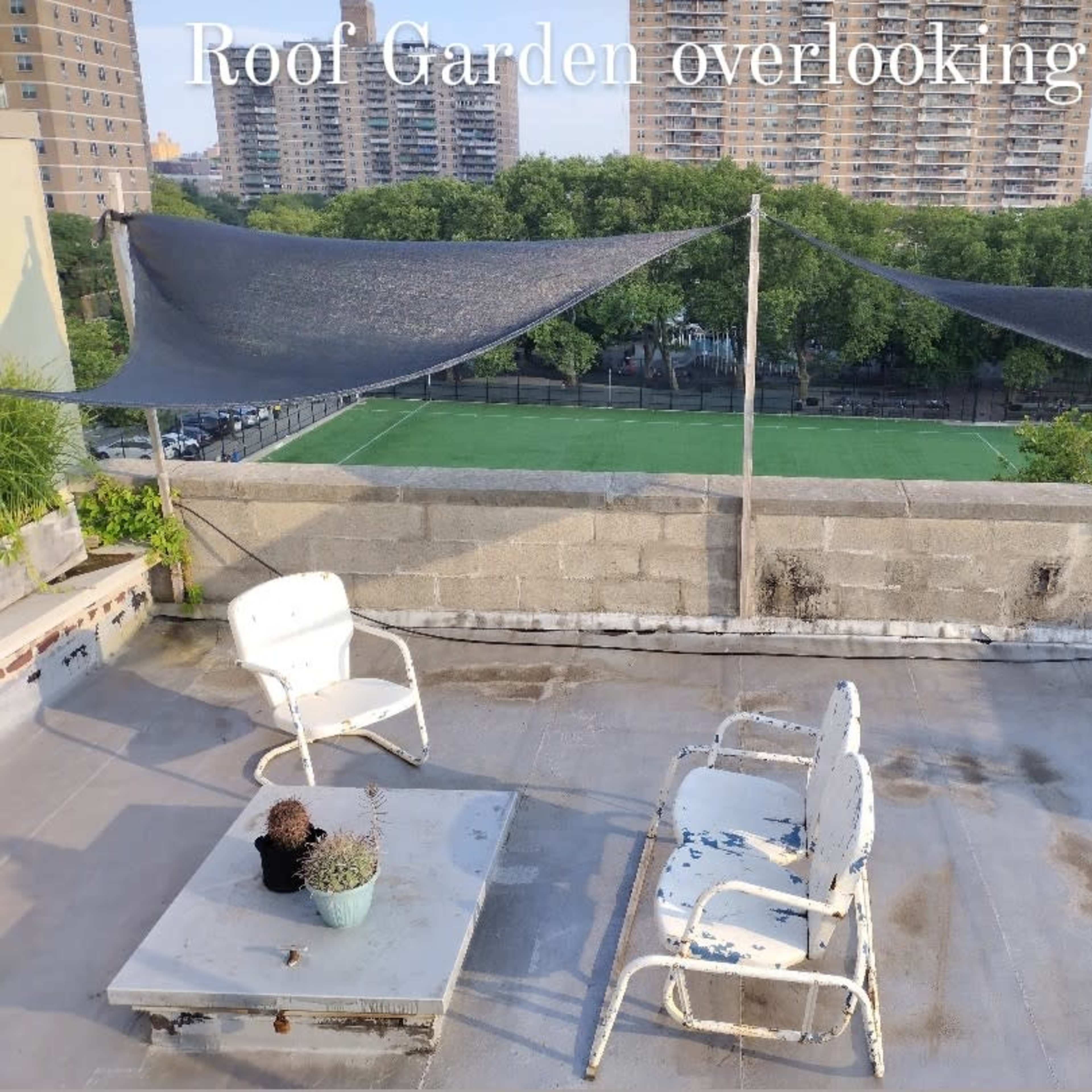 A rooftop garden with two white chairs and a small table, overlooking a park with a soccer field in the distance.