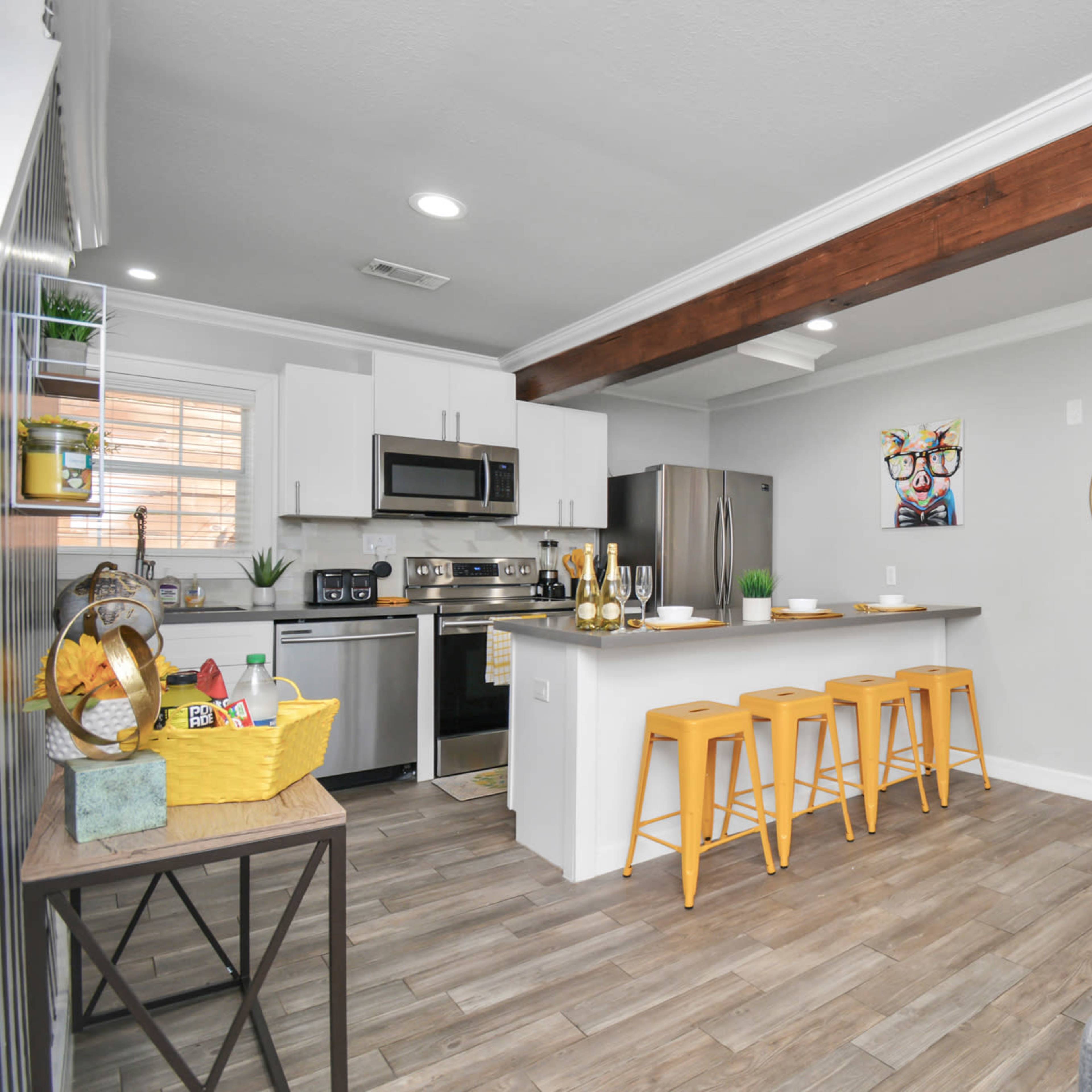 A modern kitchen and living area feature stainless steel appliances, a wooden beam ceiling, yellow bar stools, and a view into a bedroom with yellow and gray decor.