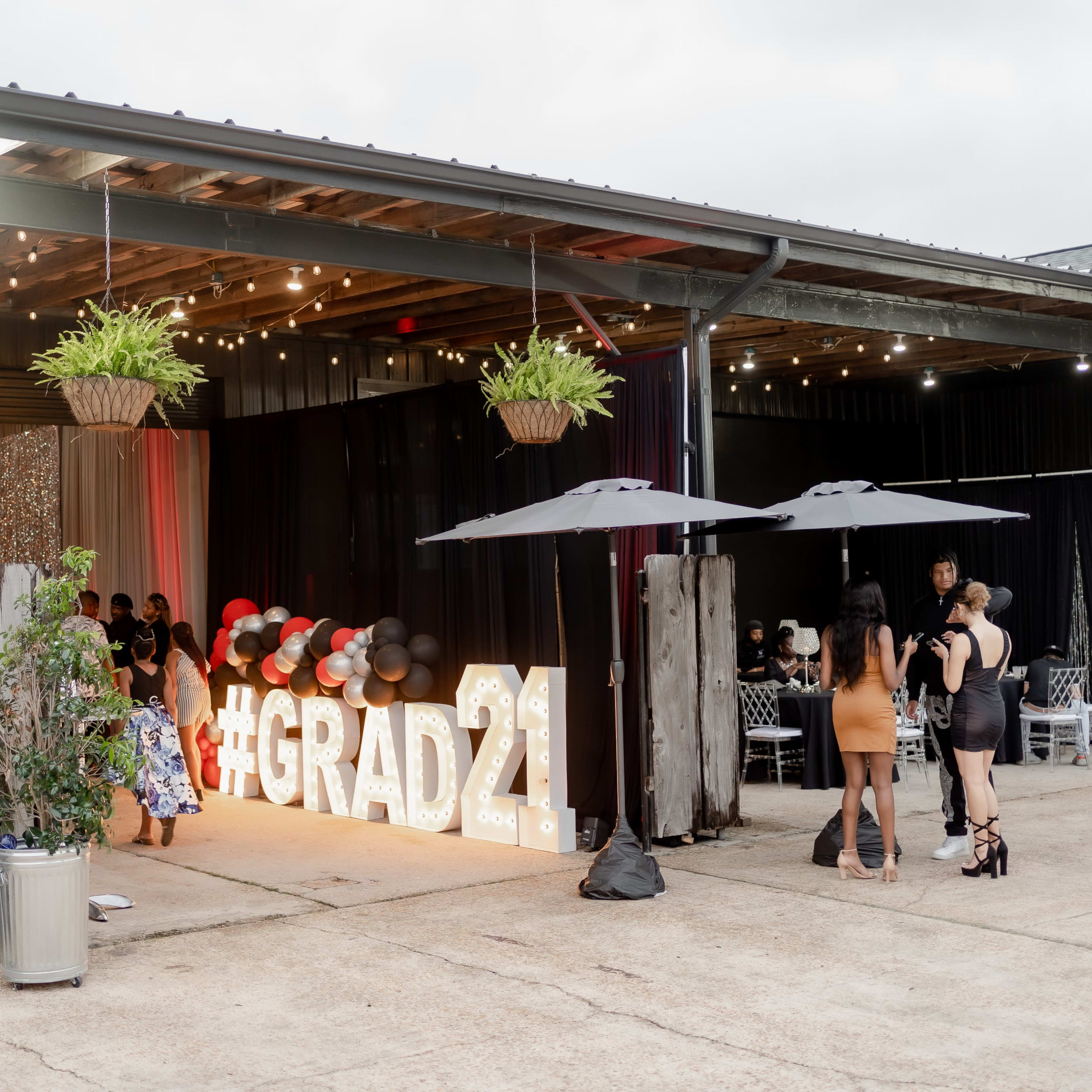 A graduation celebration venue features "#GRAD21" illuminated signage, hanging plants, and guests mingling under umbrellas.