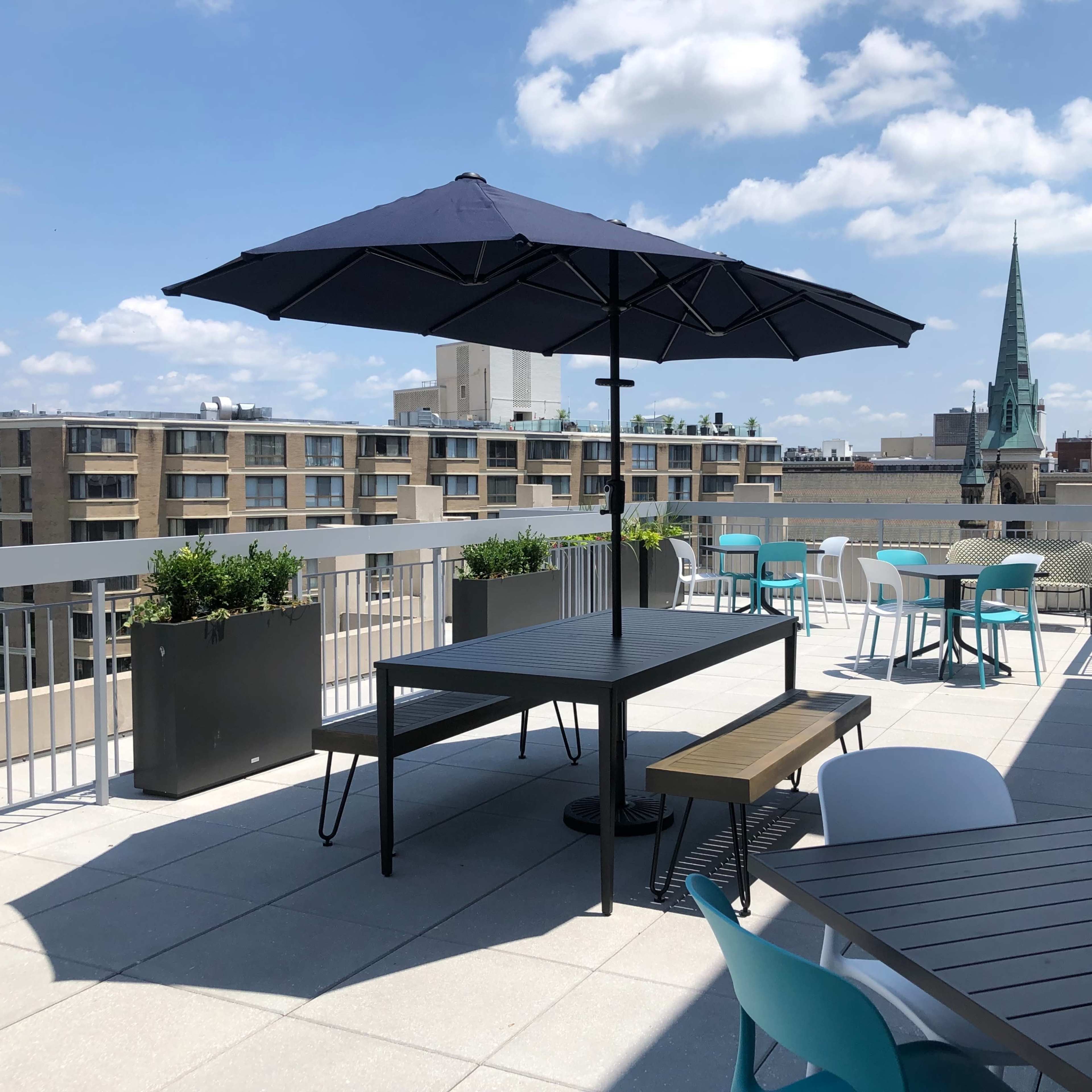 A rooftop terrace with tables, chairs, and a large umbrella, overlooking a cityscape featuring buildings and a steeple in the background.