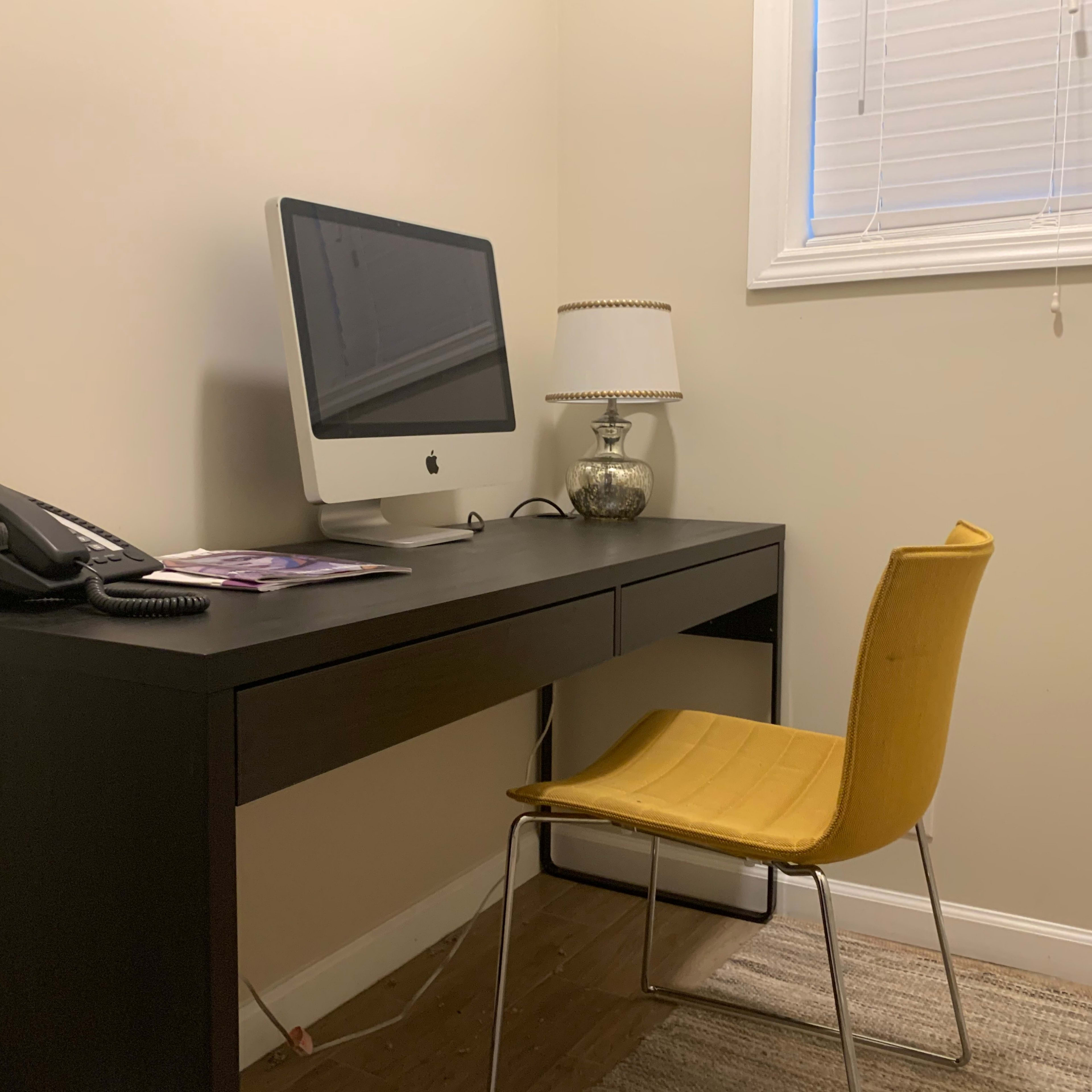 A simple workspace features an iMac on a black desk, a yellow chair, a telephone, and a lamp beside a window with closed blinds.