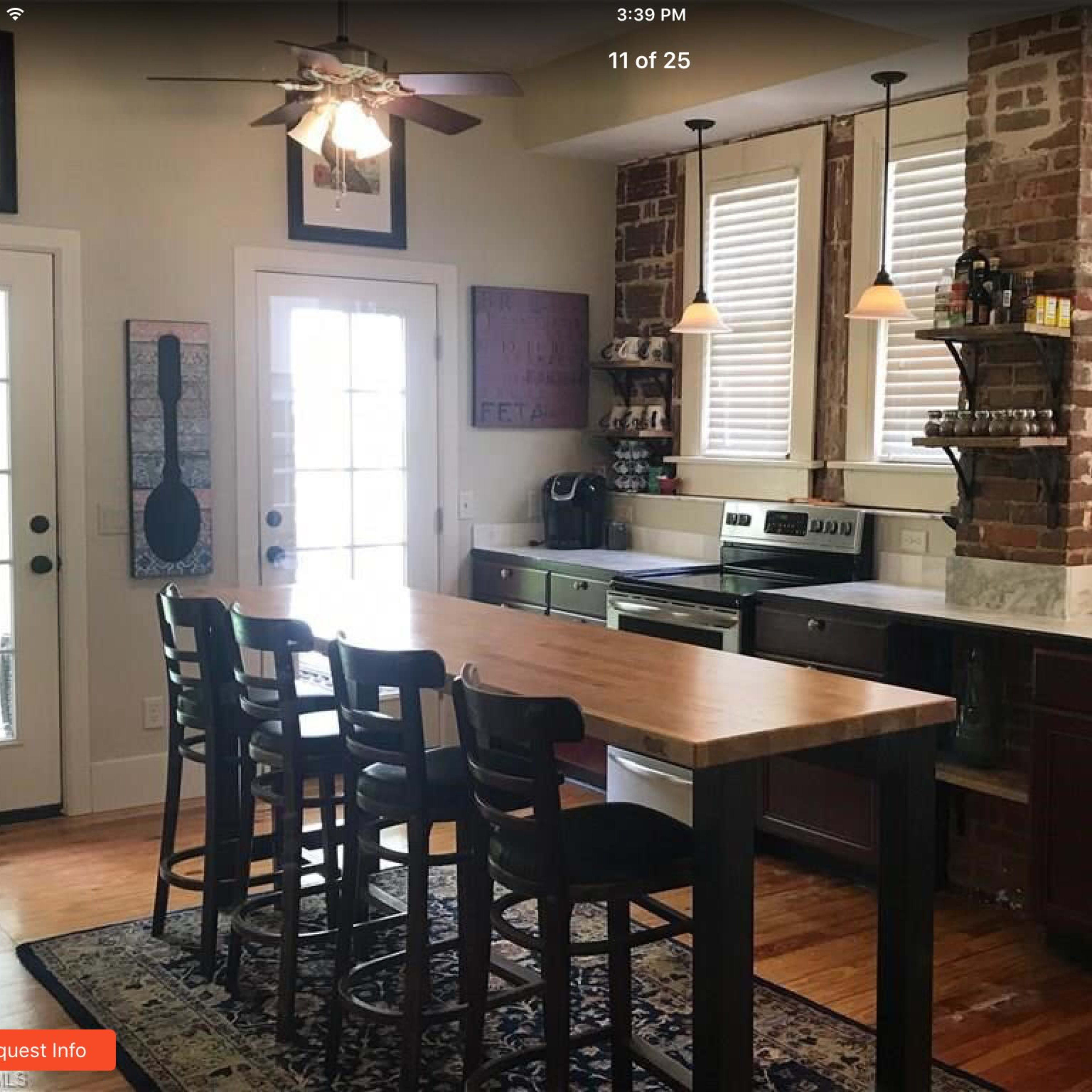A kitchen with exposed brick walls, wooden cabinetry, a central island with four bar stools, and large windows letting in natural light.