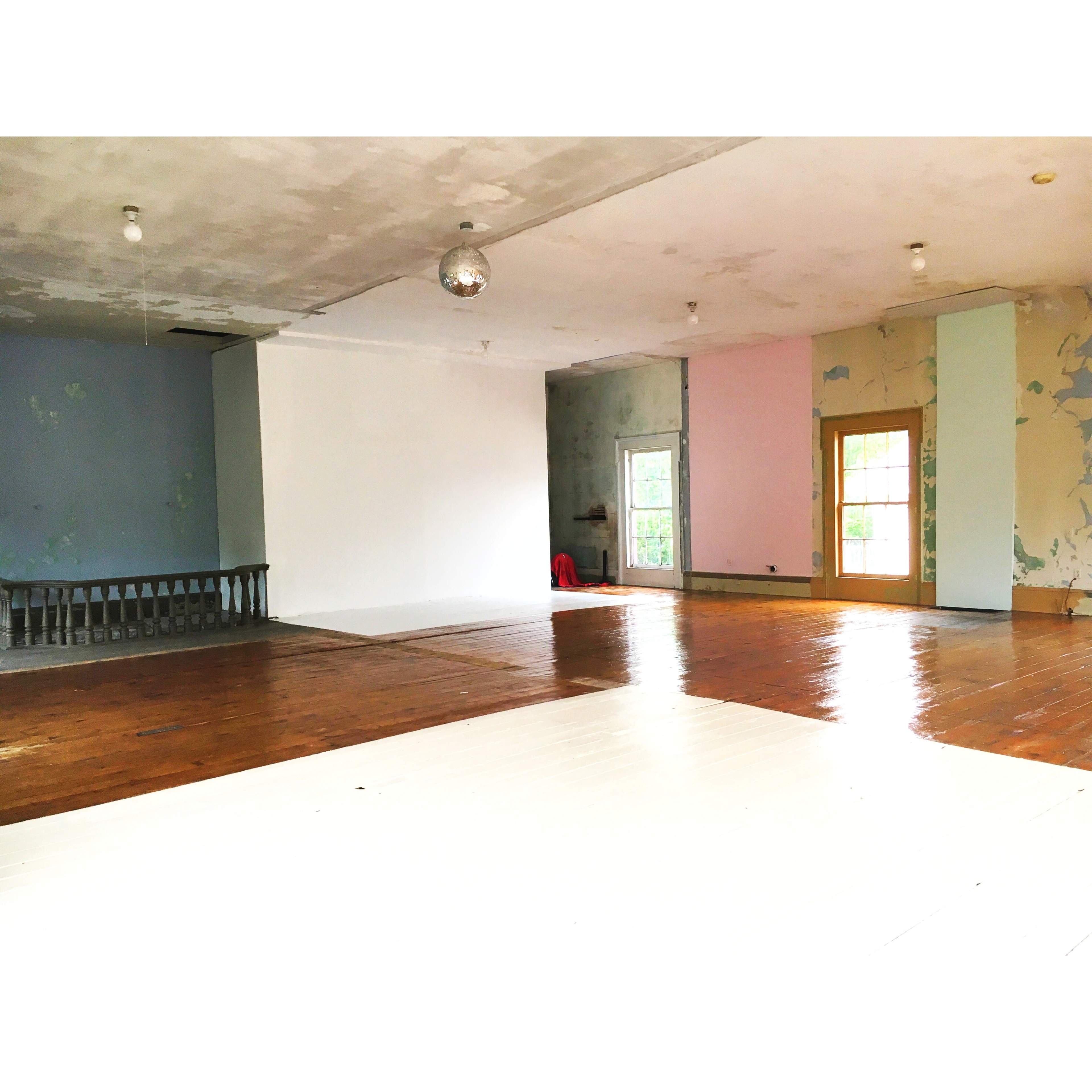 An empty interior room with a wooden floor, peeling paint on the walls, and a disco ball hanging from the ceiling.