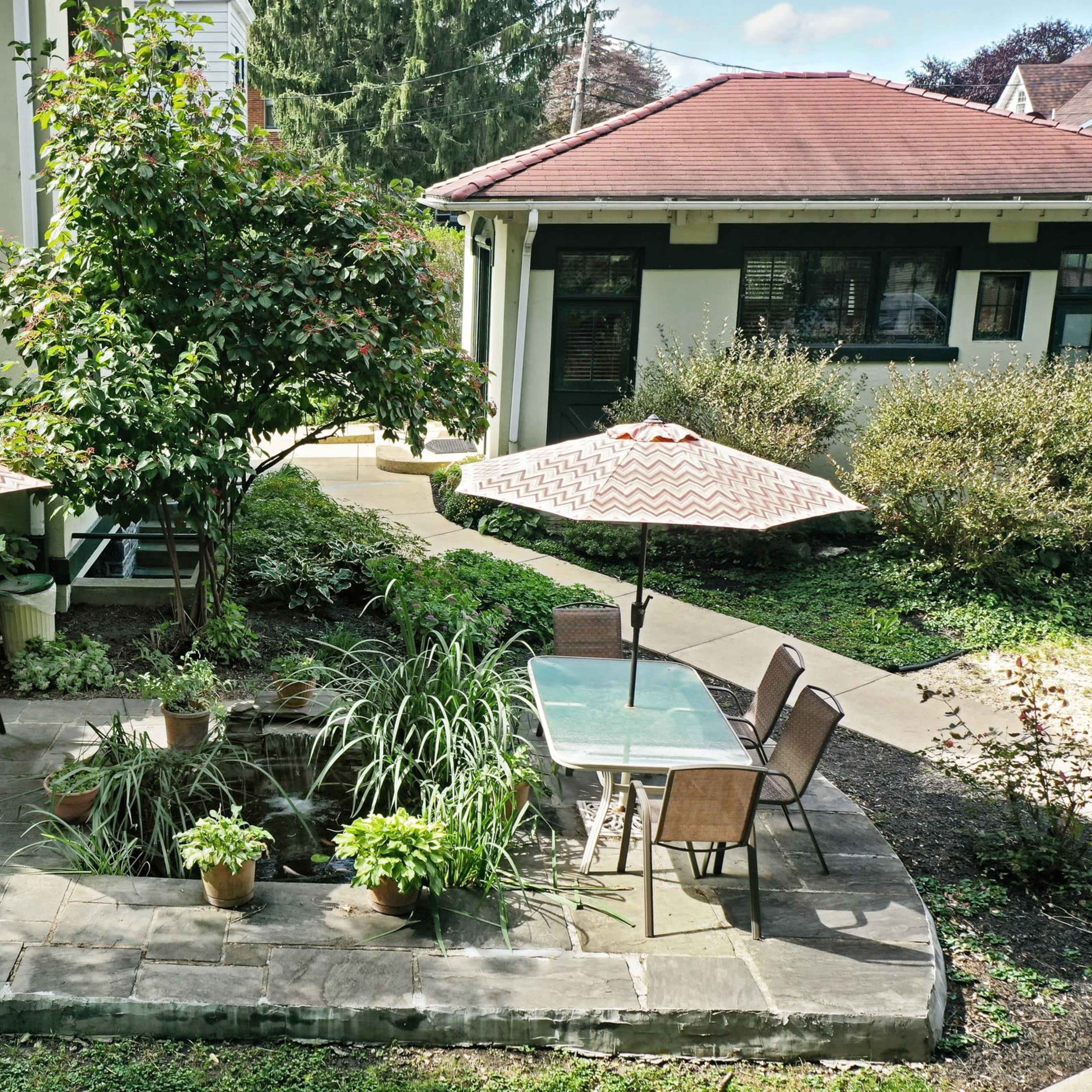 A patio featuring two shaded tables with chairs surrounded by greenery and a small pond.