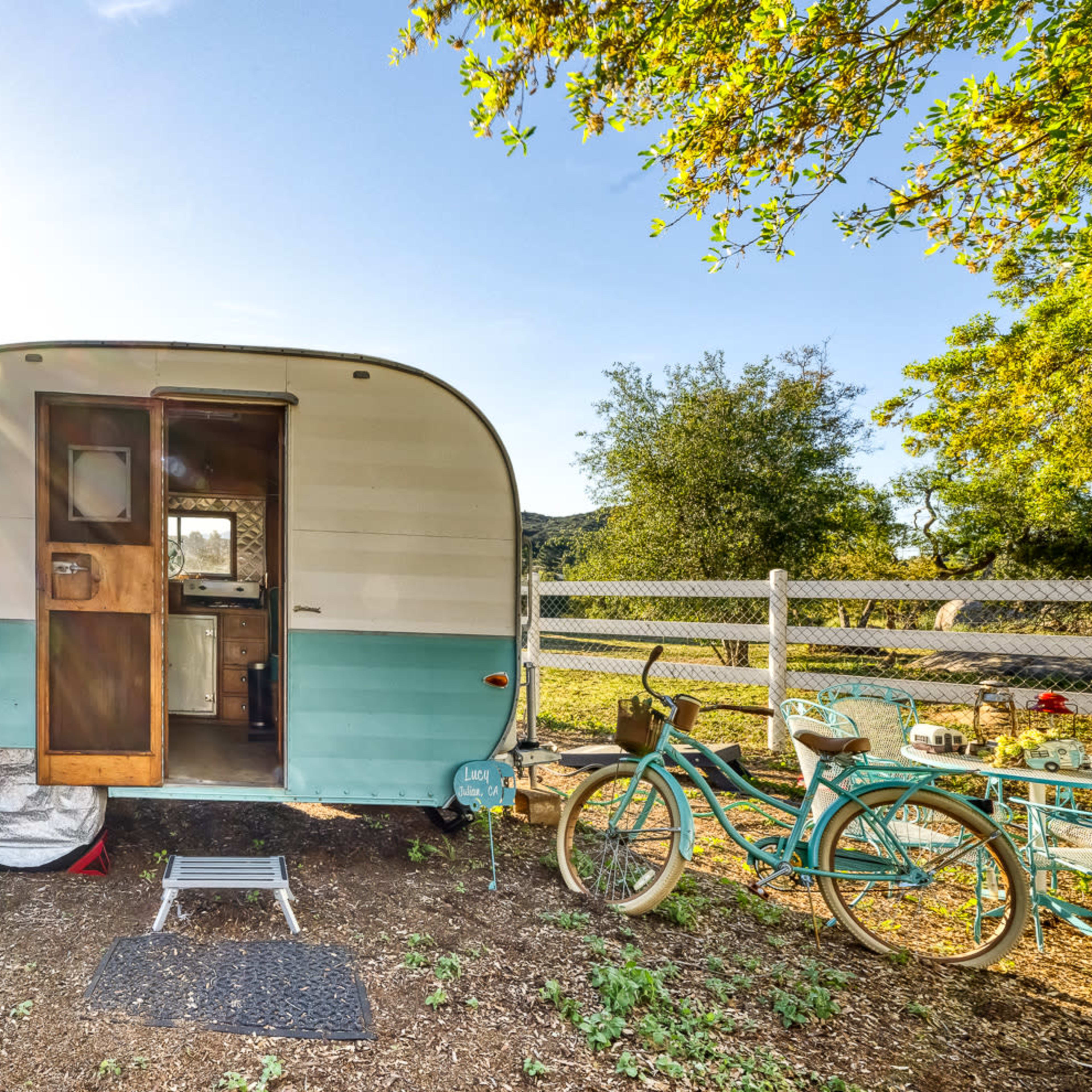A vintage camper trailer is parked in a sunlit outdoor area, with green trees and a bicycle nearby.