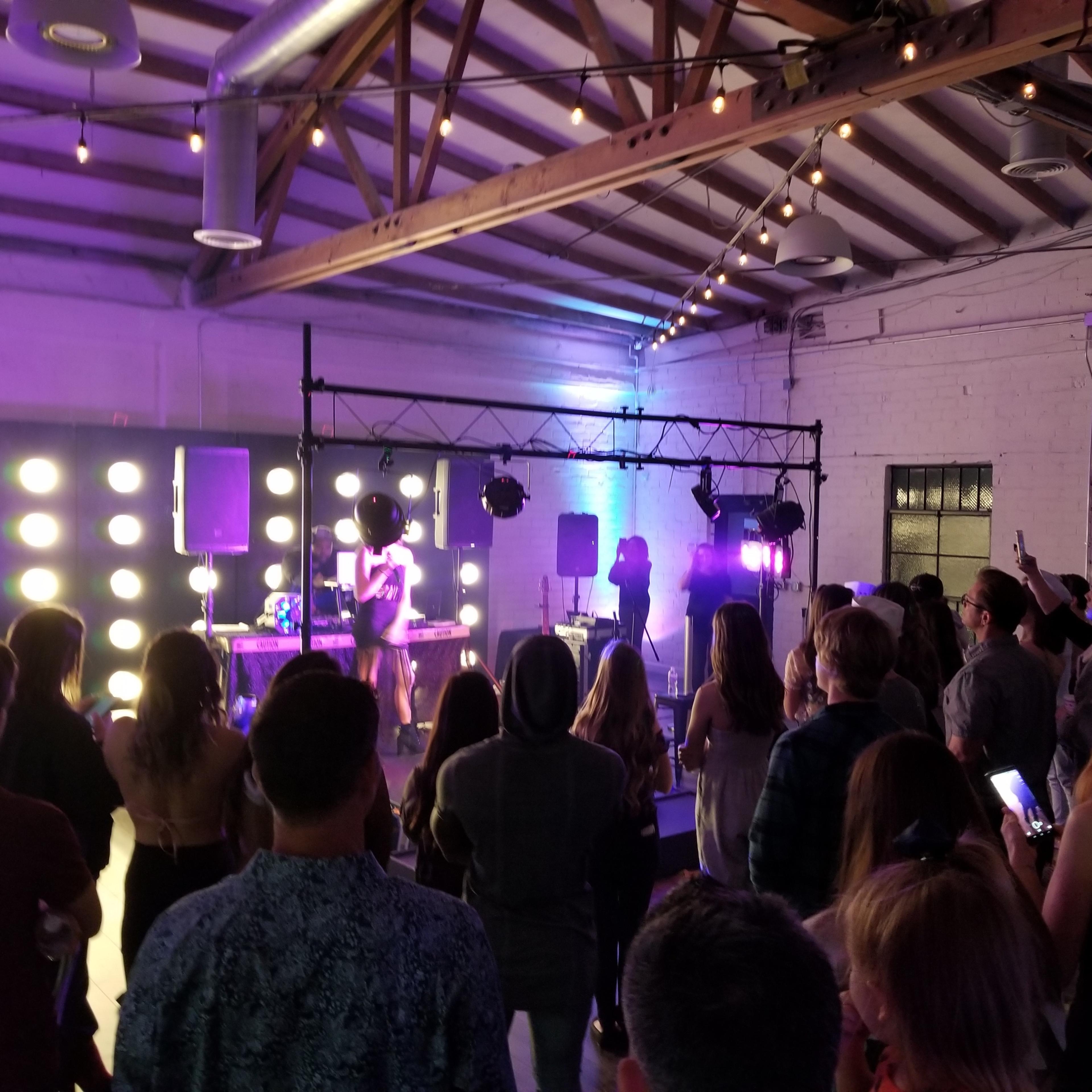 A crowd watches a performer on stage in a brightly lit venue with an industrial design.
