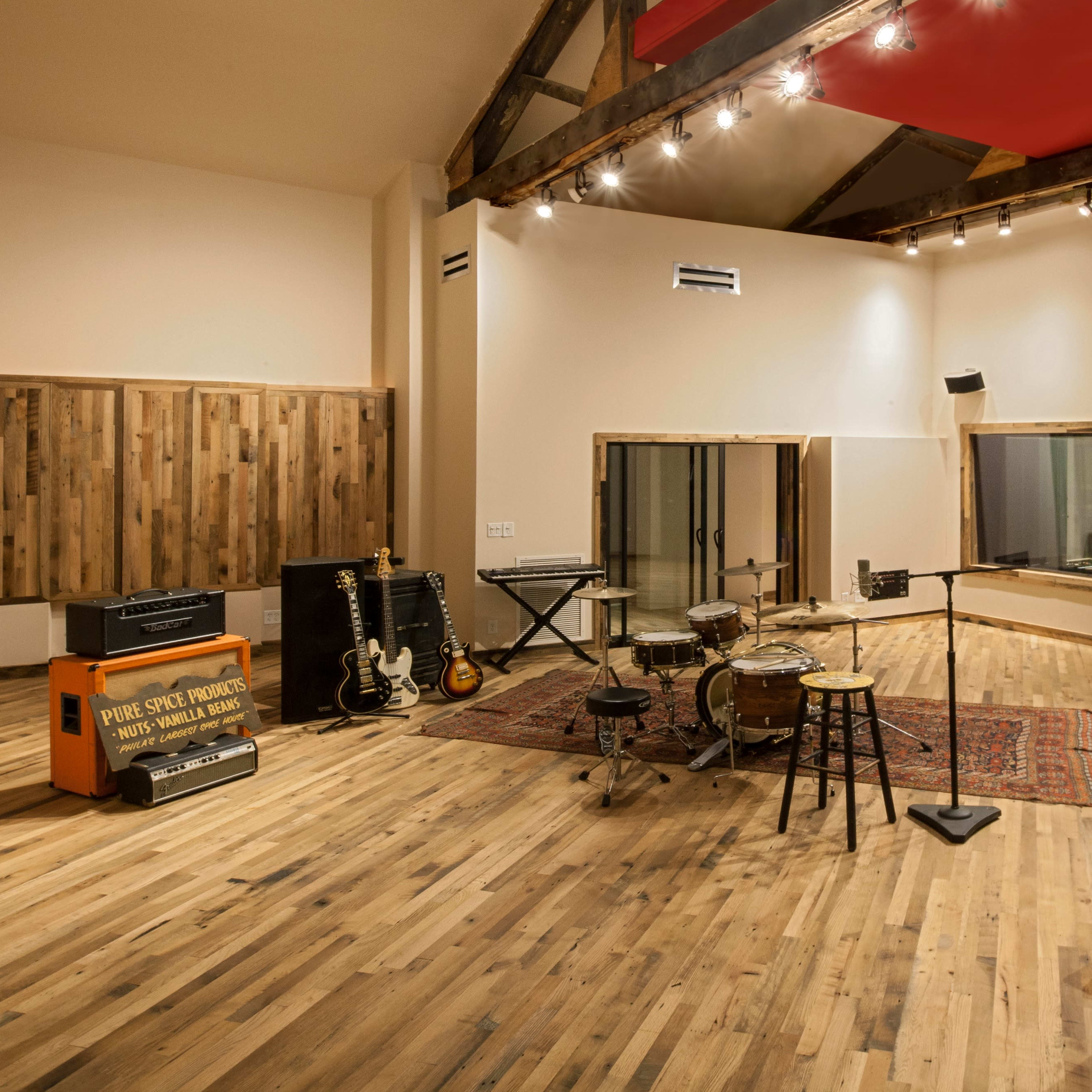 A well-lit music studio featuring various instruments, including guitars, a drum set, and sound equipment, arranged in a spacious wooden interior.