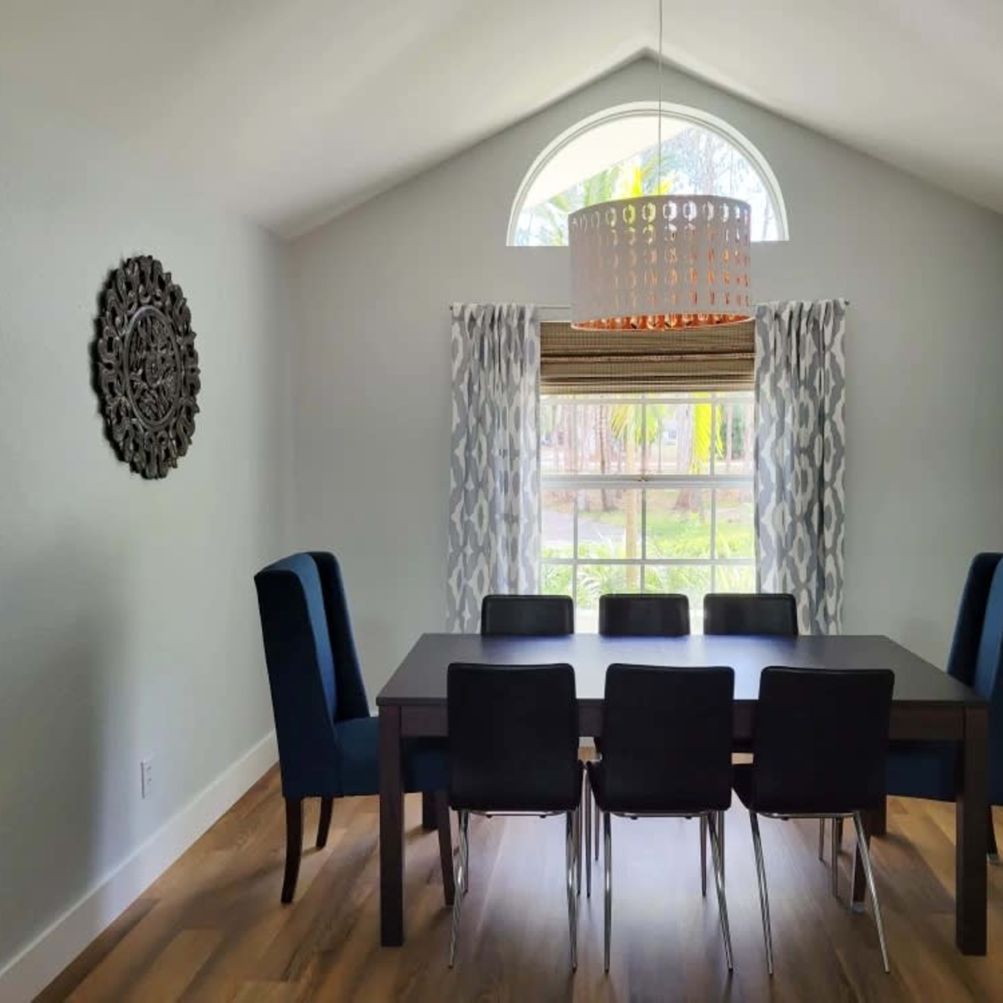 A modern dining room features a rectangular table surrounded by black chairs and blue upholstered armchairs, with a large window and decorative lighting above.