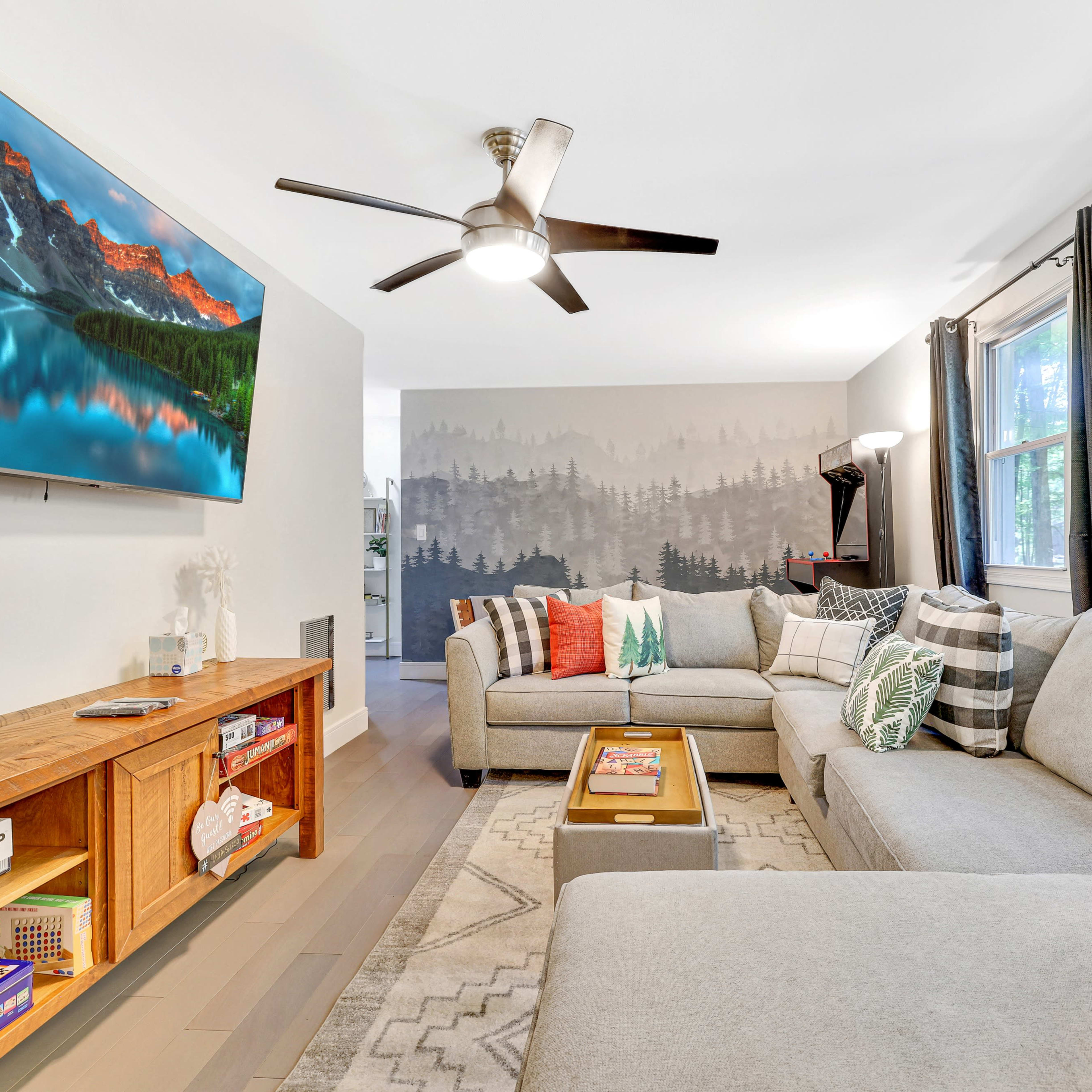A living room with a sectional sofa, a wooden coffee table, a television on the wall, and large windows allowing natural light to enter.