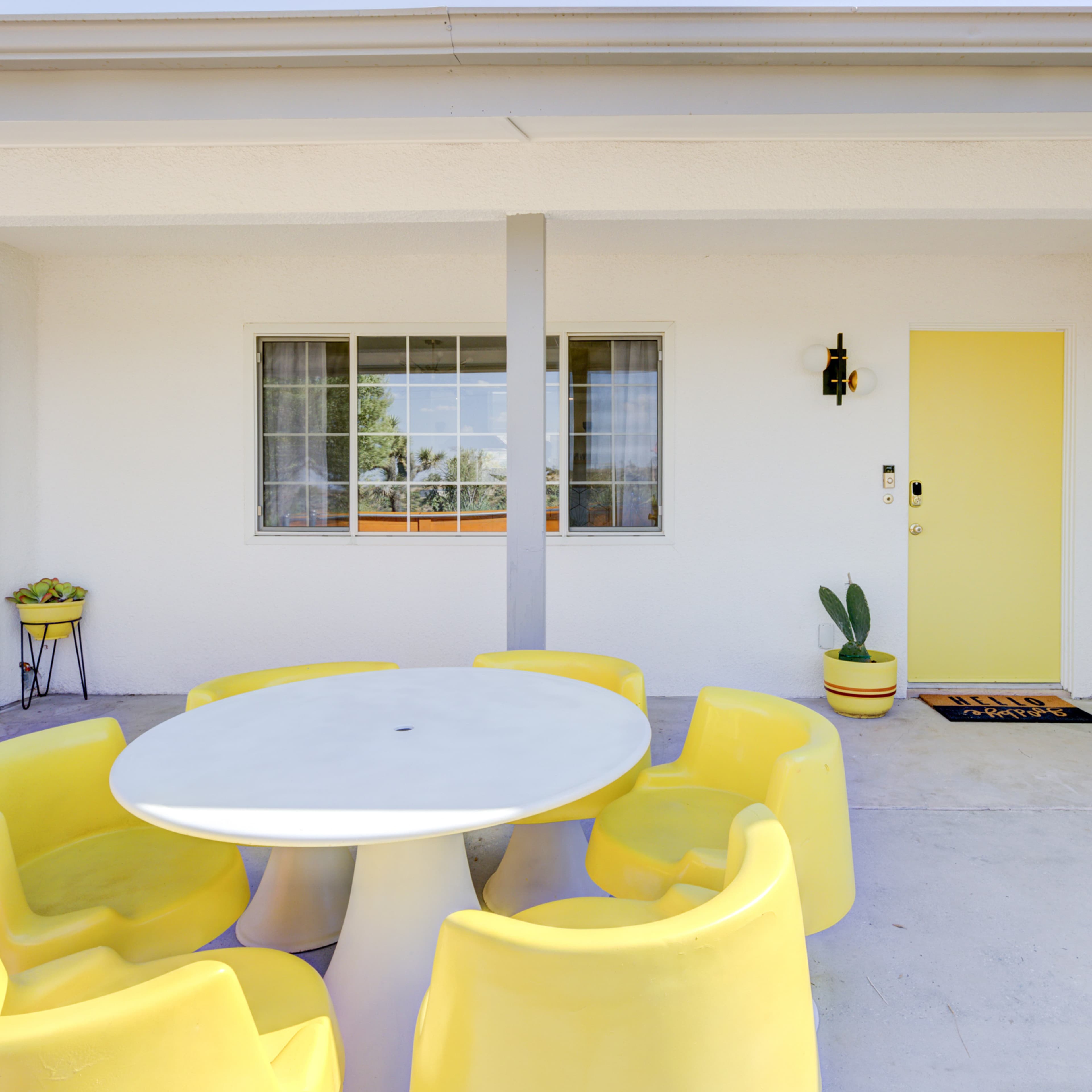 A porch with a round white table surrounded by six yellow chairs, alongside a door painted yellow and potted plants.
