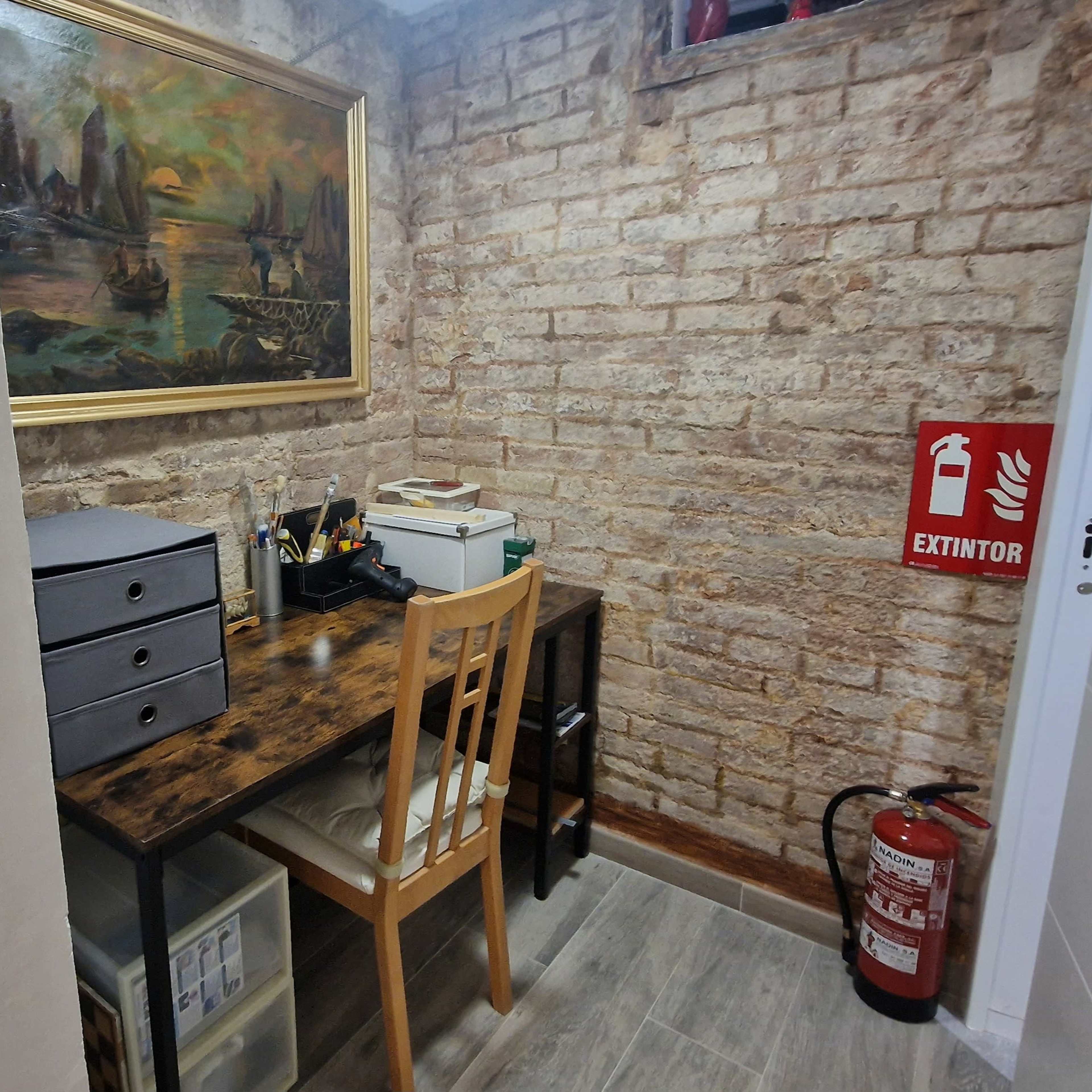 A small office space with a wooden desk and a chair against a brick wall, featuring a painting on the wall and a fire extinguisher nearby.
