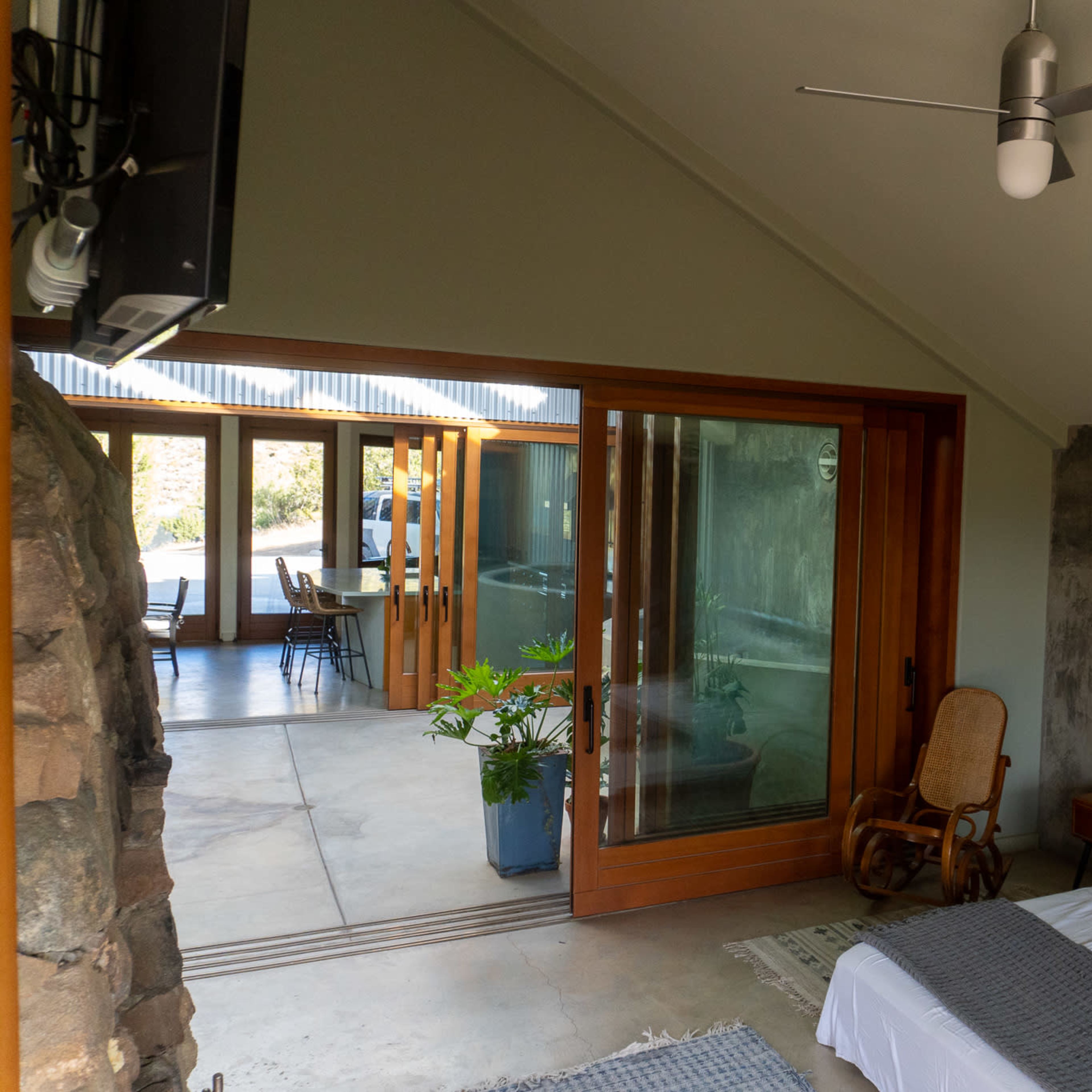 A bedroom with a bed and a wooden rocking chair, leading to a glass sliding door that opens to a bright living area.